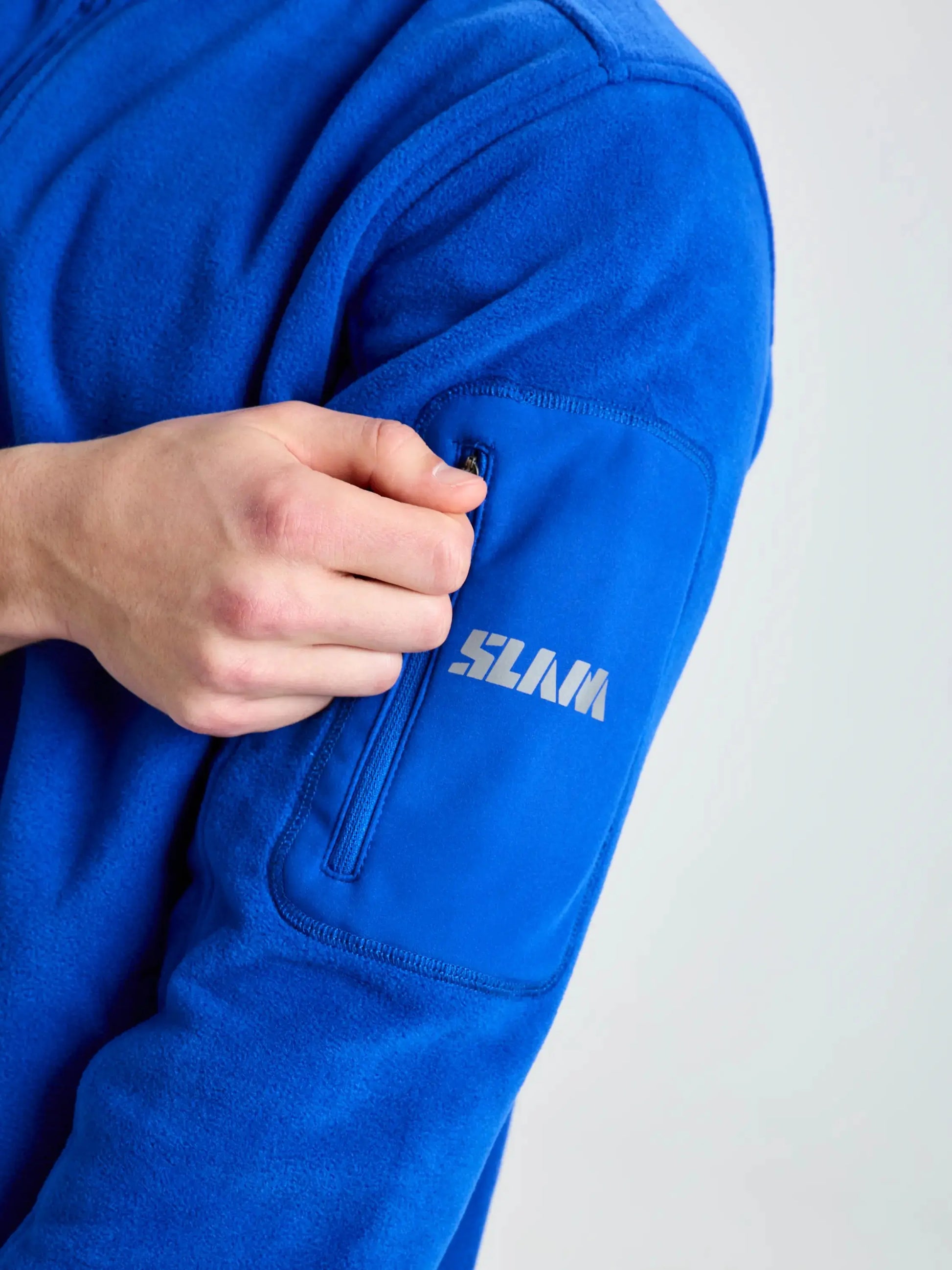 A person wearing a vibrant blue men’s DECK FLEECE jacket by SLAM opens a small zippered pocket on the upper left sleeve, where the brand name “SLAM” is printed in white near the zipper. Made from recycled microfibre fleece, this heavyweight jacket stands out against the plain, light-colored background.