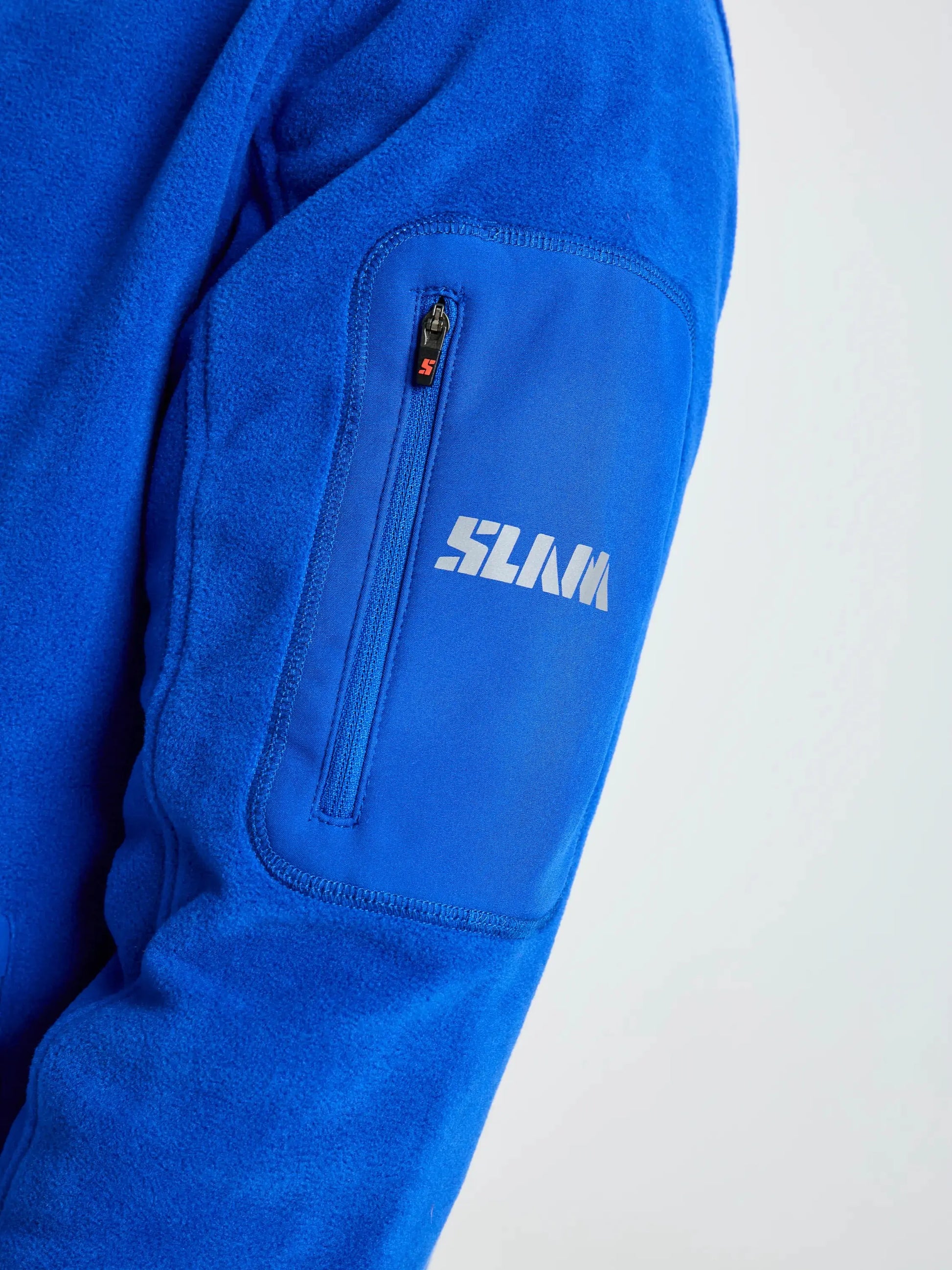 A close-up of the sleeve of the SLAM DECK FLEECE jacket, made from blue recycled microfibre fleece, featuring a zippered pocket with “SLAM” printed in white on it. The background is a plain, light color.