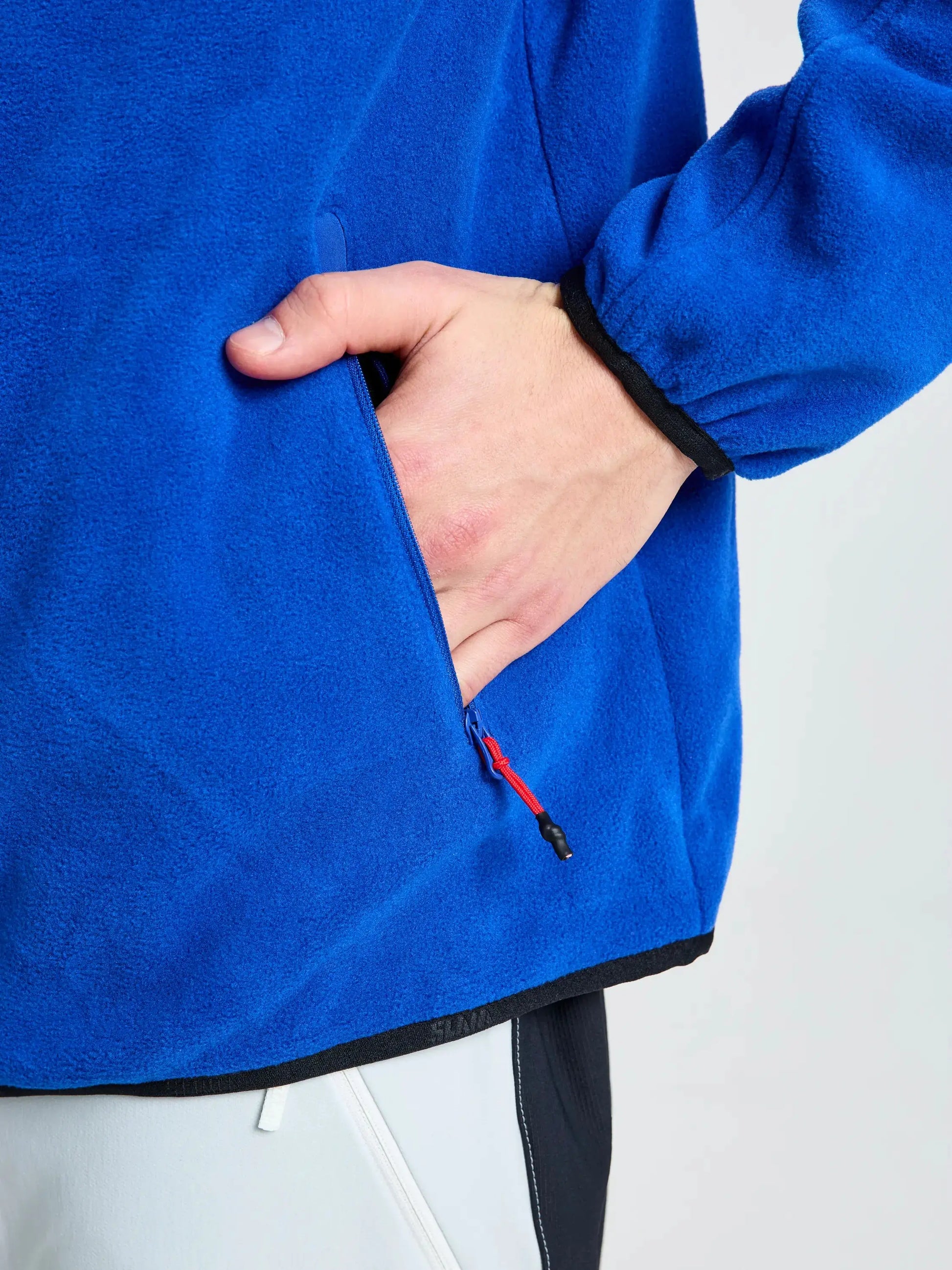 A person wearing the SLAM DECK FLEECE men’s blue jacket with black trim places their right hand in a zippered side pocket. The heavy fleece material provides warmth, and the bottom of their white and black pants is partially visible.