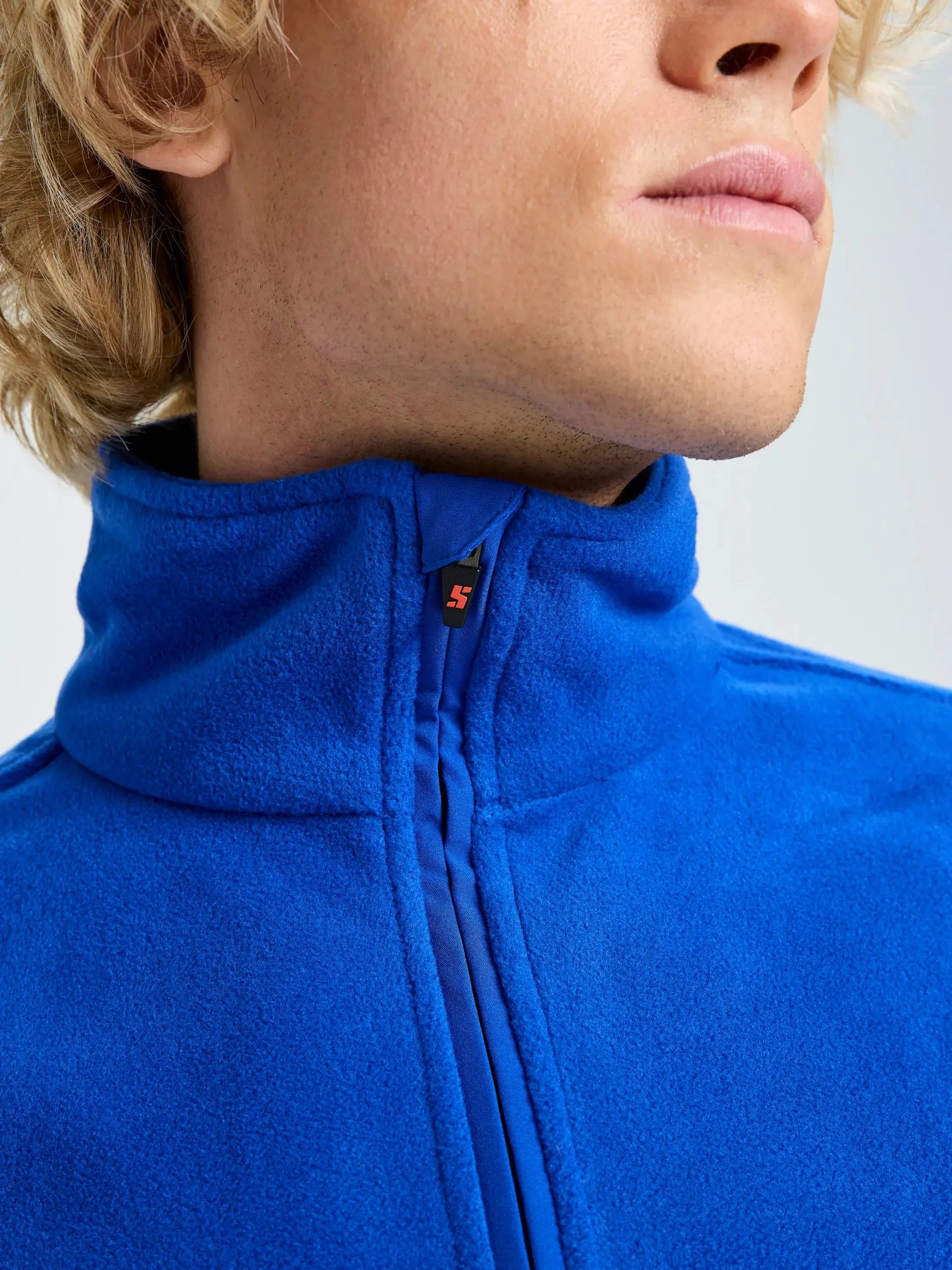 Close-up of a person wearing the SLAM DECK FLEECE, a bright blue recycled microfibre jacket zipped up to the collar. The lower part of their face and neck, with light skin and blond hair, are visible against a plain light background.