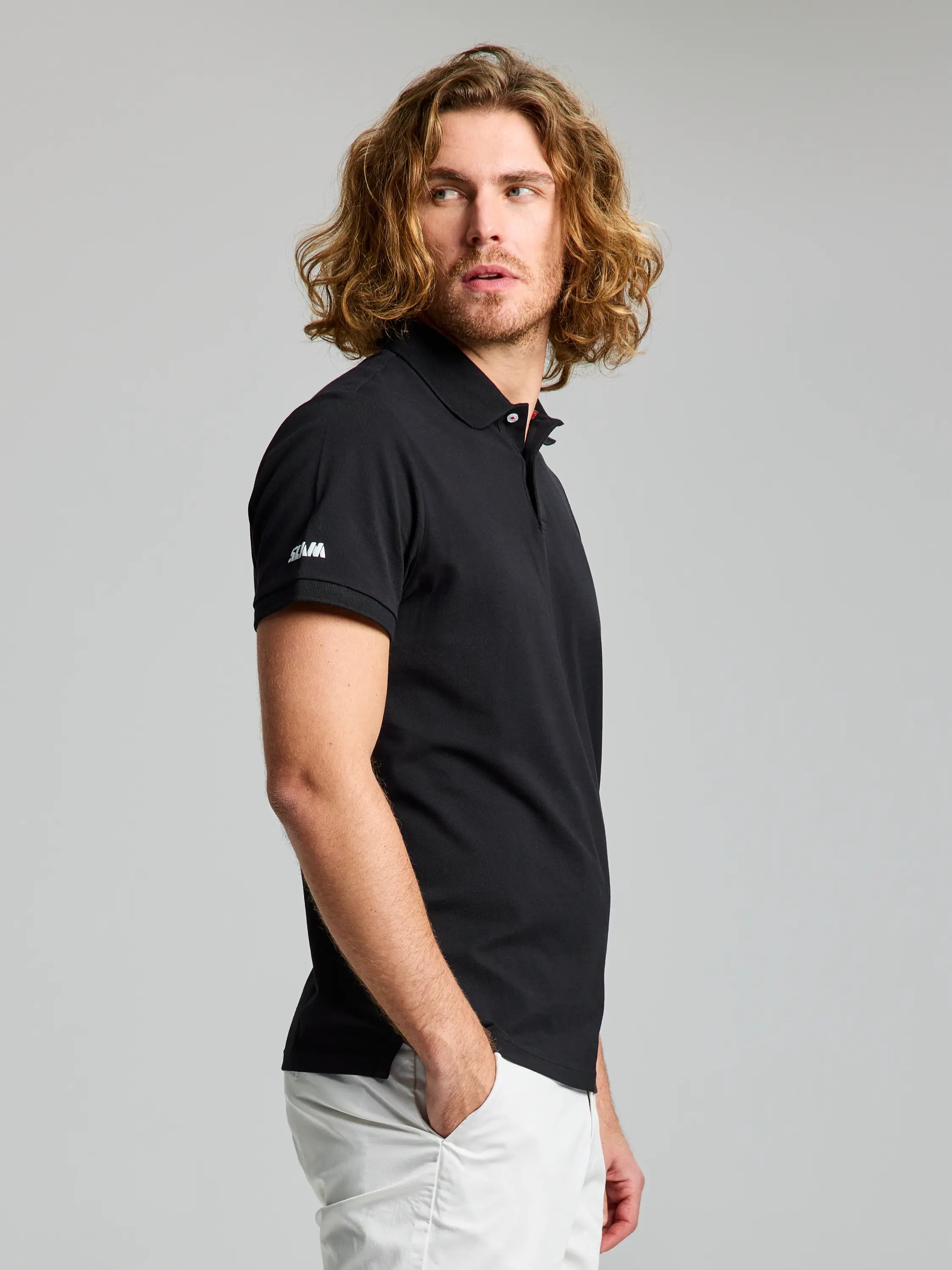 A person with long, wavy hair is wearing the DECK POLO SHORT SLEEVE from SLAM, a black men's shirt crafted from breathable cotton pique, paired with white pants. They are standing against a plain background, looking to the side with one hand in their pocket. The shirt features a logo on the sleeve.
