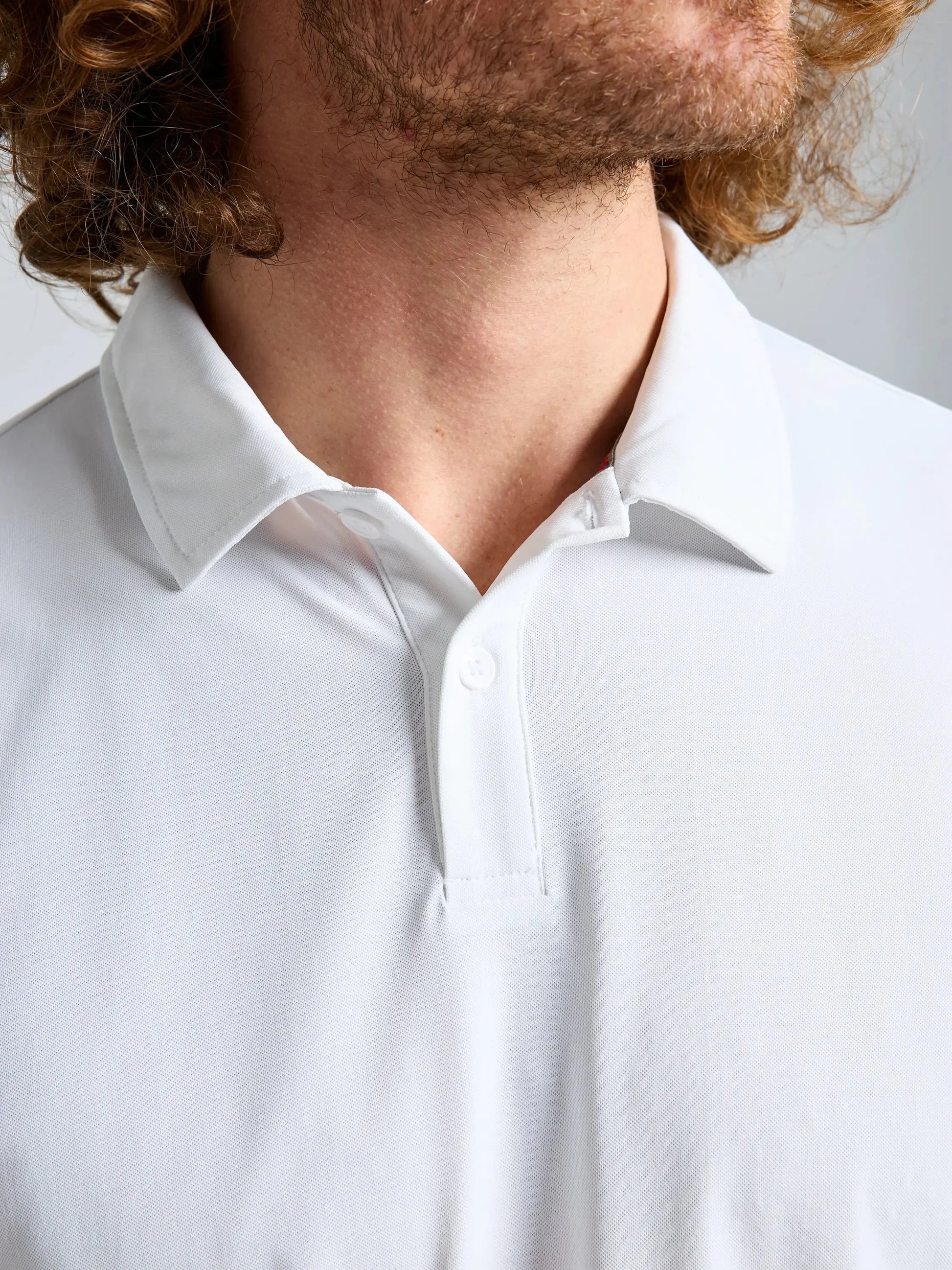 Close-up of a person wearing the SLAM TECH PIQUE POLO SS in white, featuring UVP 50+ protection and one button fastened. Only the lower face, neck, and some curly brown hair are visible; the rest of the face is partially out of frame.