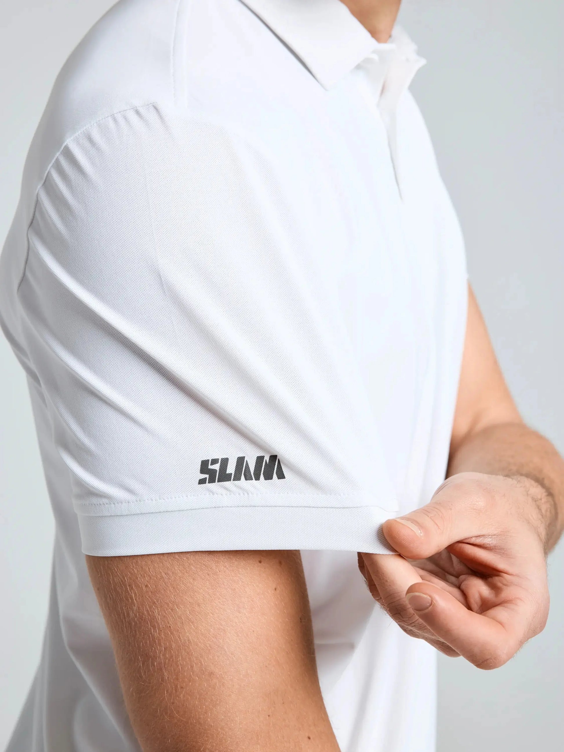 A person wearing the SLAM TECH PIQUE POLO SS in white holds the sleeve’s edge, revealing a black SLAM logo printed near the hem. Only their torso and part of one arm are visible.