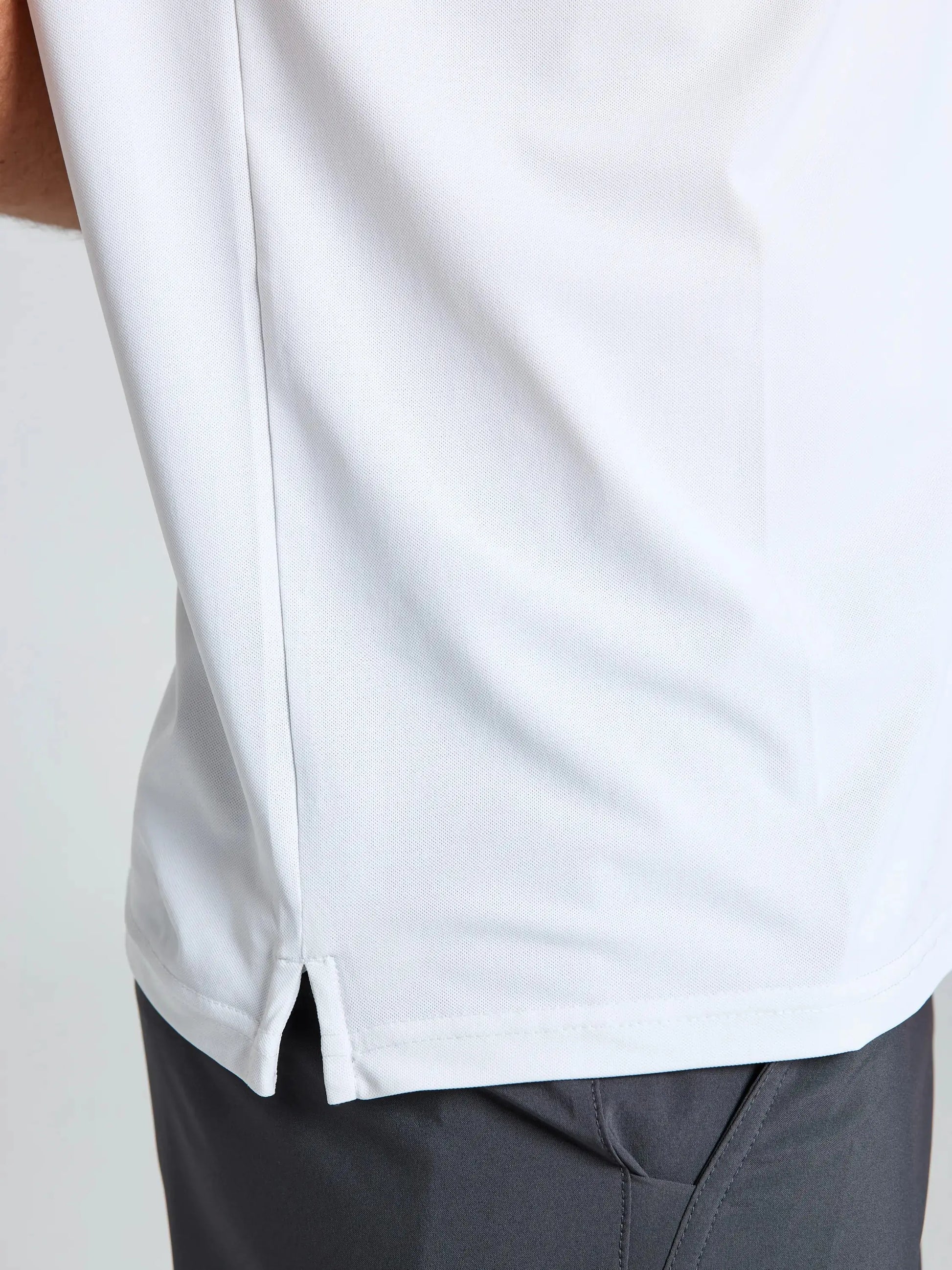 Close-up of a person wearing the SLAM TECH PIQUE POLO SS in white, featuring short sleeves and a small side slit at the hem, paired with dark gray pants. Only part of the torso and upper legs are shown against a plain, light-colored background.
