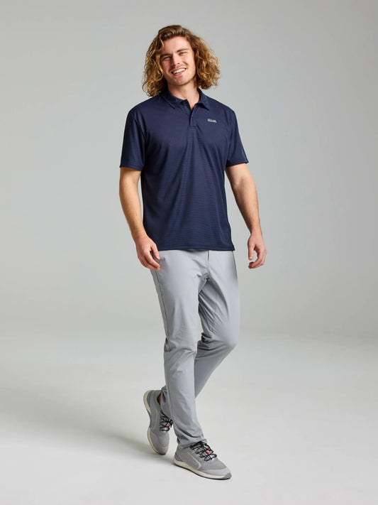 A person with long curly hair is wearing an ACTIVE QD POLO by SLAM, along with gray pants and sneakers, standing against a plain gray background. They are smiling and have a relaxed posture.