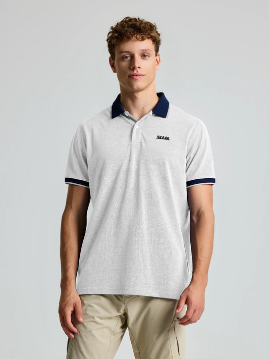 A man with short curly hair wears the SLAM POLO HEBA EVO SS—a light gray piqué polo with dark blue collar and sleeve trim, plus a small "SLAM" logo on the left chest—paired with light khaki pants, standing against a plain light gray background.