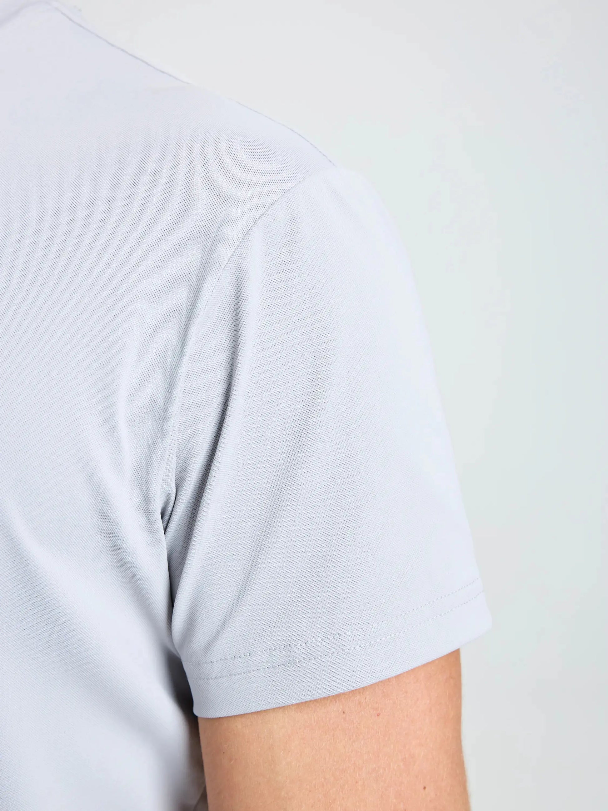 Close-up of a person’s upper arm and shoulder wearing the SLAM TECH PIQUE T-SHIRT in light gray. The shirt, designed with breathable fabric for all-day comfort, is a men’s short-sleeved style. The background is a neutral light color, showing only part of the wearer’s back and arm.