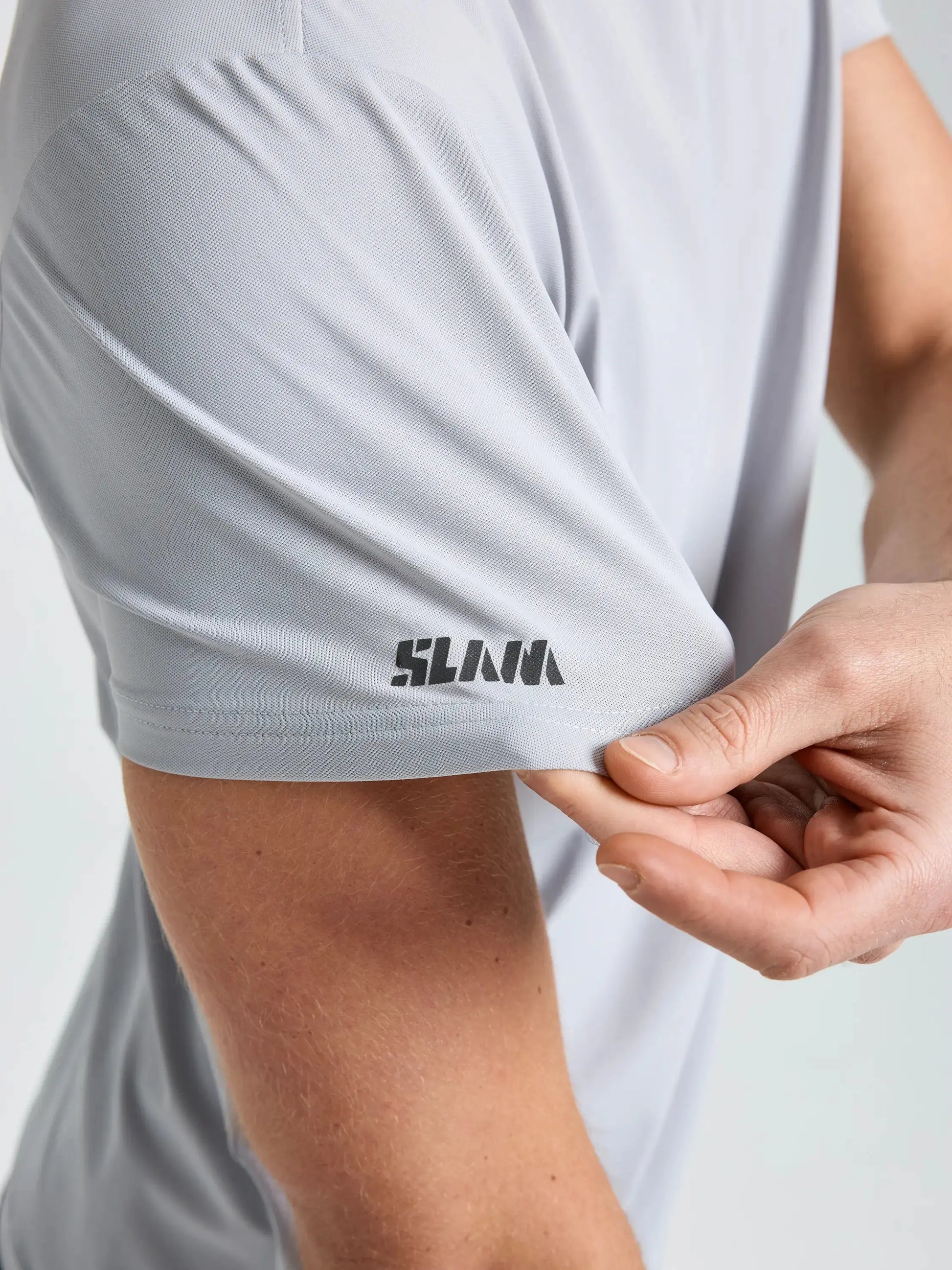 A person pulls the sleeve of a light gray SLAM TECH PIQUE T-SHIRT, revealing a black SLAM logo printed near the hem. The breathable men’s short-sleeved shirt has a close fit, and the background is plain and light-colored.