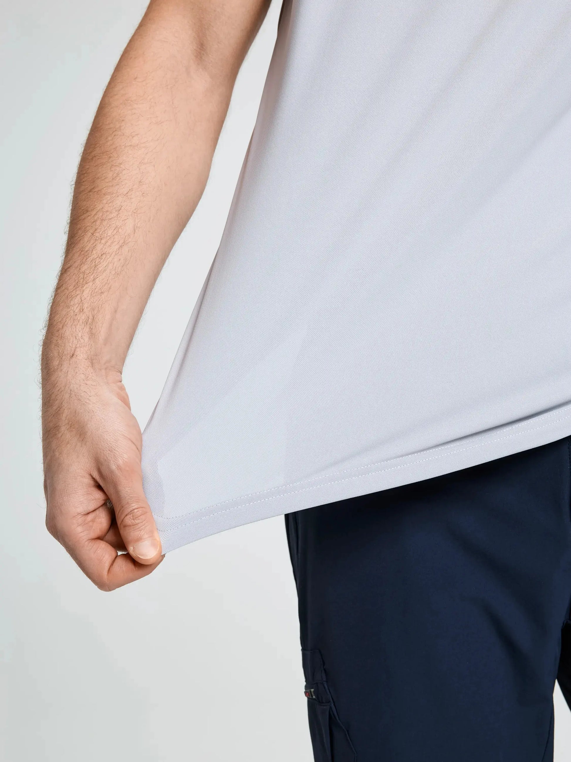 A person is shown wearing the SLAM TECH PIQUE T-SHIRT in light gray and dark blue pants, stretching the fabric outward with one hand to demonstrate its flexibility. The image focuses on the torso and part of the arm, emphasizing the breathable and comfortable design of this men’s short-sleeved T-shirt.