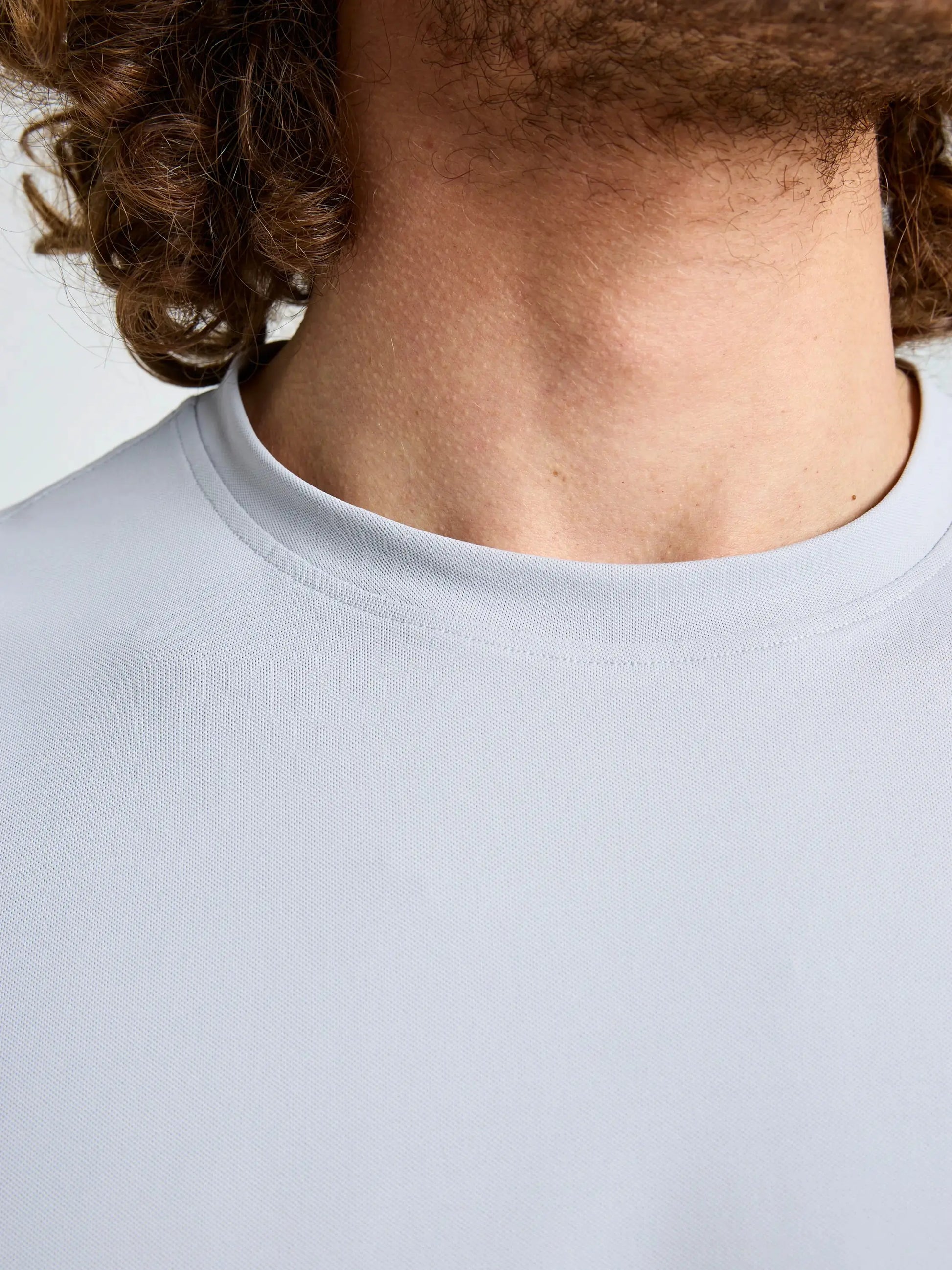 Close-up of a person’s neck and lower face with curly hair, wearing the SLAM TECH PIQUE T-SHIRT—a men’s short-sleeved crew neck tee in plain light grey. Made from breathable, UV-resistant fabric, this shirt stands out against a simple, light-colored background.