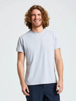 A person with curly, shoulder-length hair is standing and smiling. They are wearing the SLAM TECH PIQUE T-SHIRT in light gray, a breathable men’s short-sleeved top, paired with dark pants. The background is plain and light-colored.