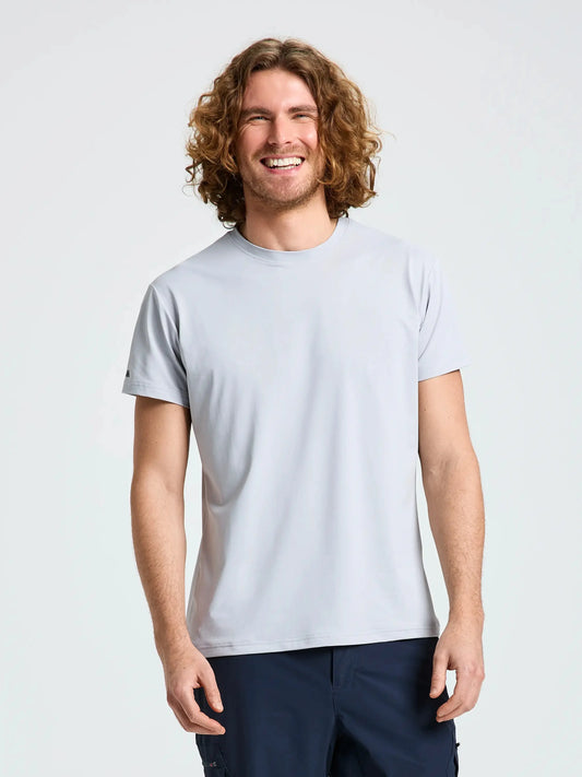 A person with curly, shoulder-length hair is standing and smiling. They are wearing the SLAM TECH PIQUE T-SHIRT in light gray, a breathable men’s short-sleeved top, paired with dark pants. The background is plain and light-colored.