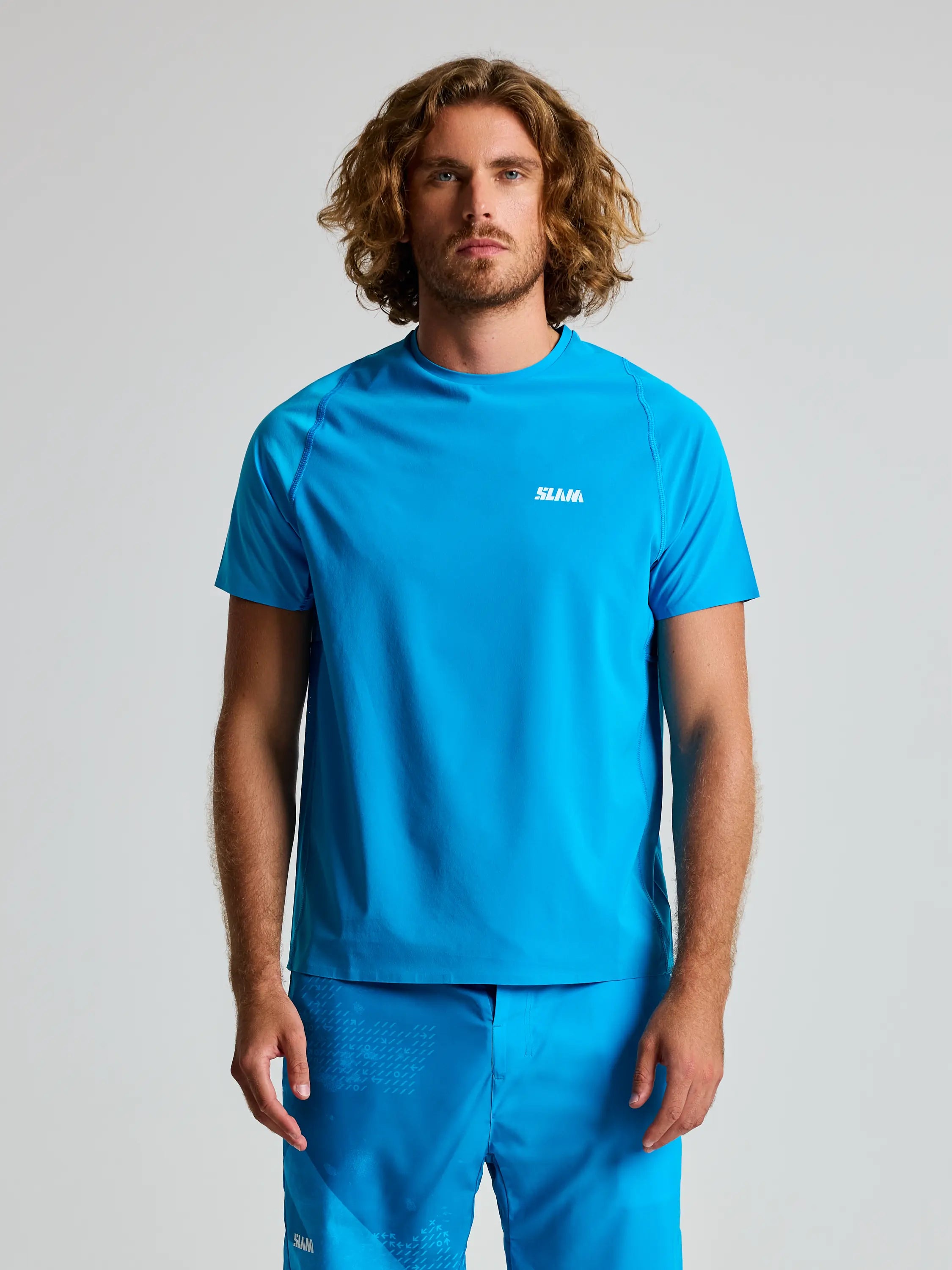 A man with long curly hair models a matching blue athletic shirt and shorts featuring "SLAM" boldly on his chest. The OD TECH T-SHIRT SS offers UV protection, ideal for active days, against a plain gray background.