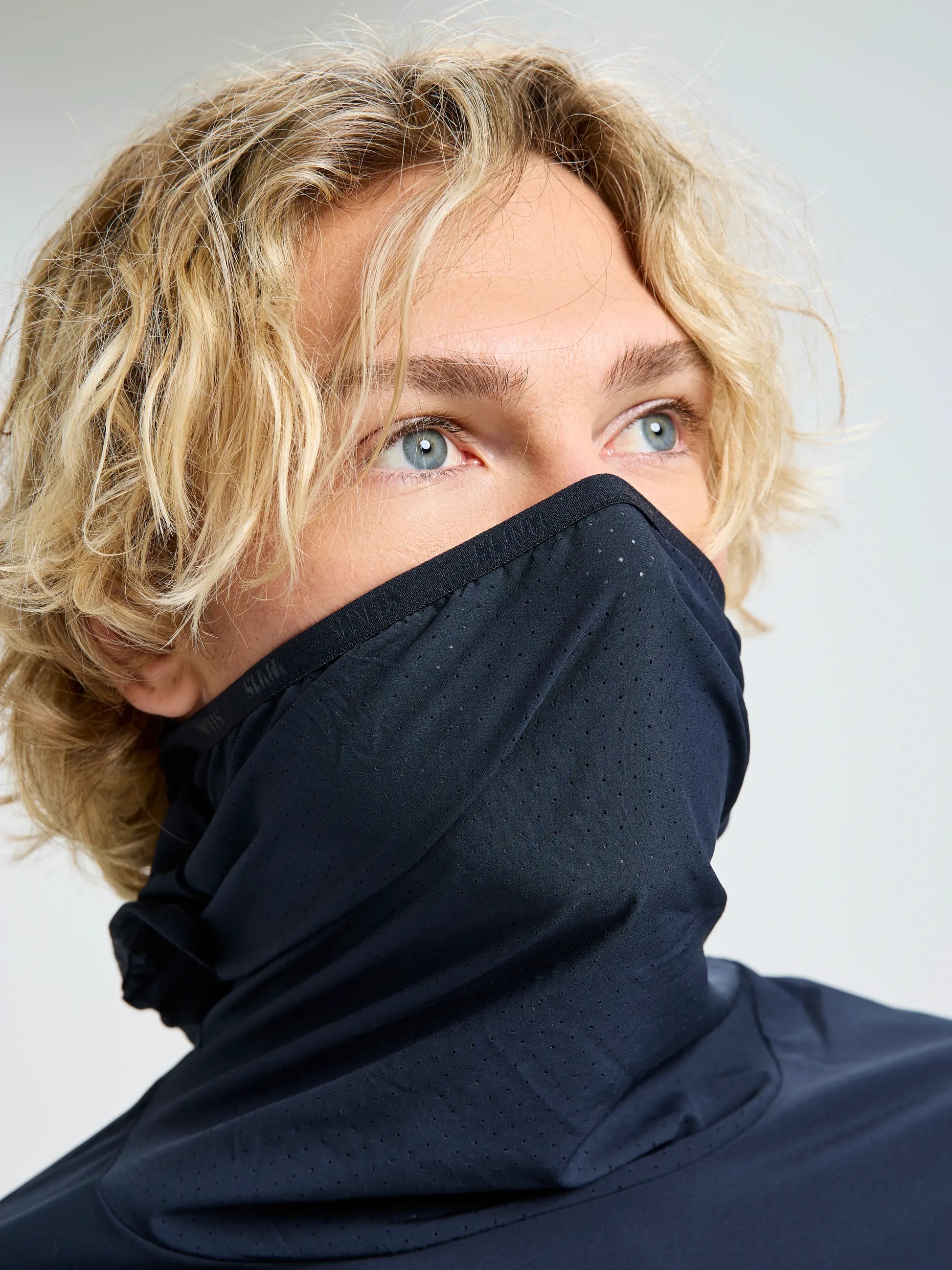 A person with wavy blond hair and blue eyes looks to the side, wearing a navy blue OD TECH HOODED T-SHIRT by SLAM and a matching perforated face covering, both made from recycled stretch fabric.