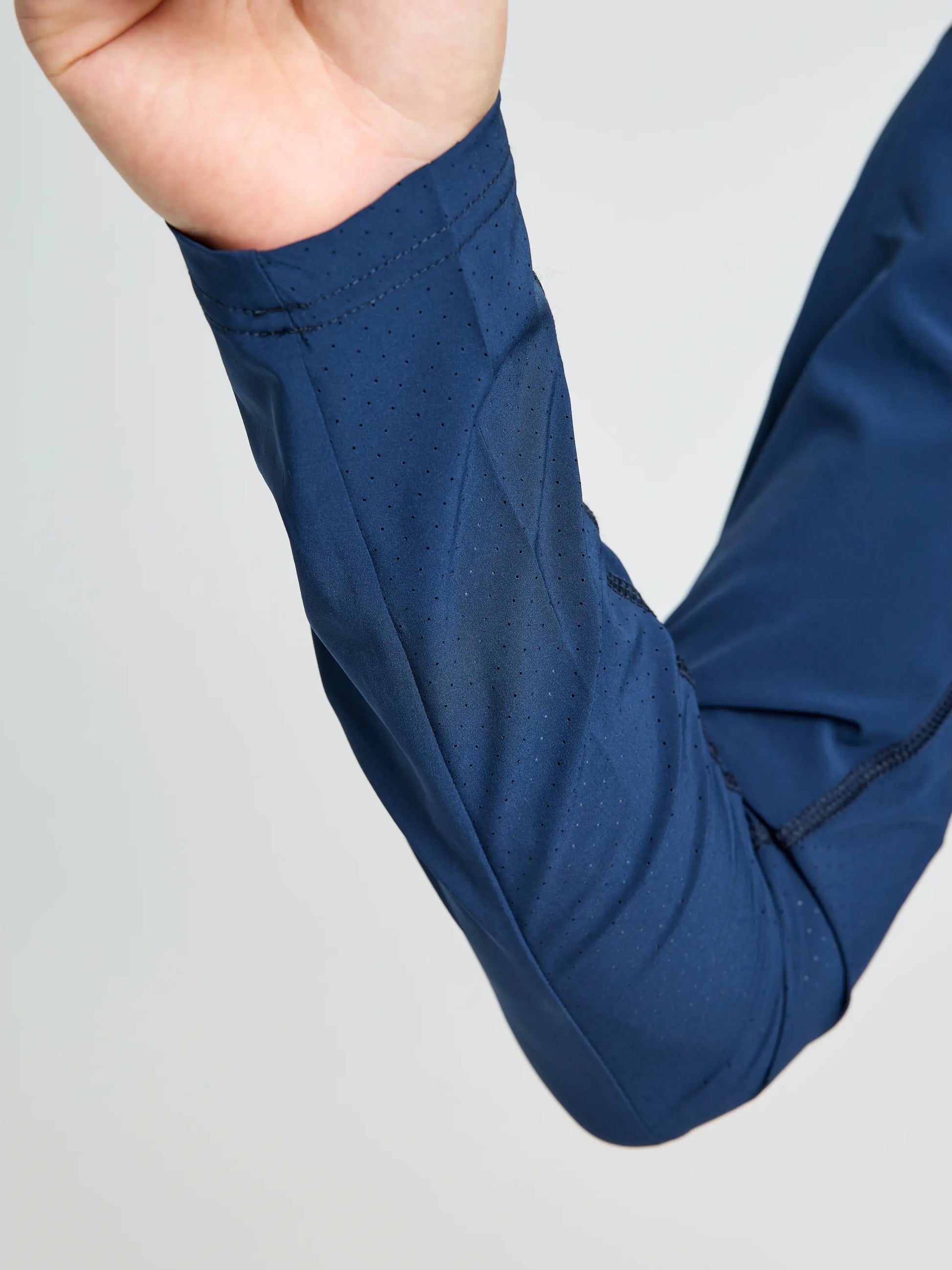 A close-up shows a bent arm in the SLAM OD TECH HOODED T-SHIRT, a long-sleeved dark blue top made from recycled stretch fabric with visible ventilation perforations. The background is plain and light-colored.