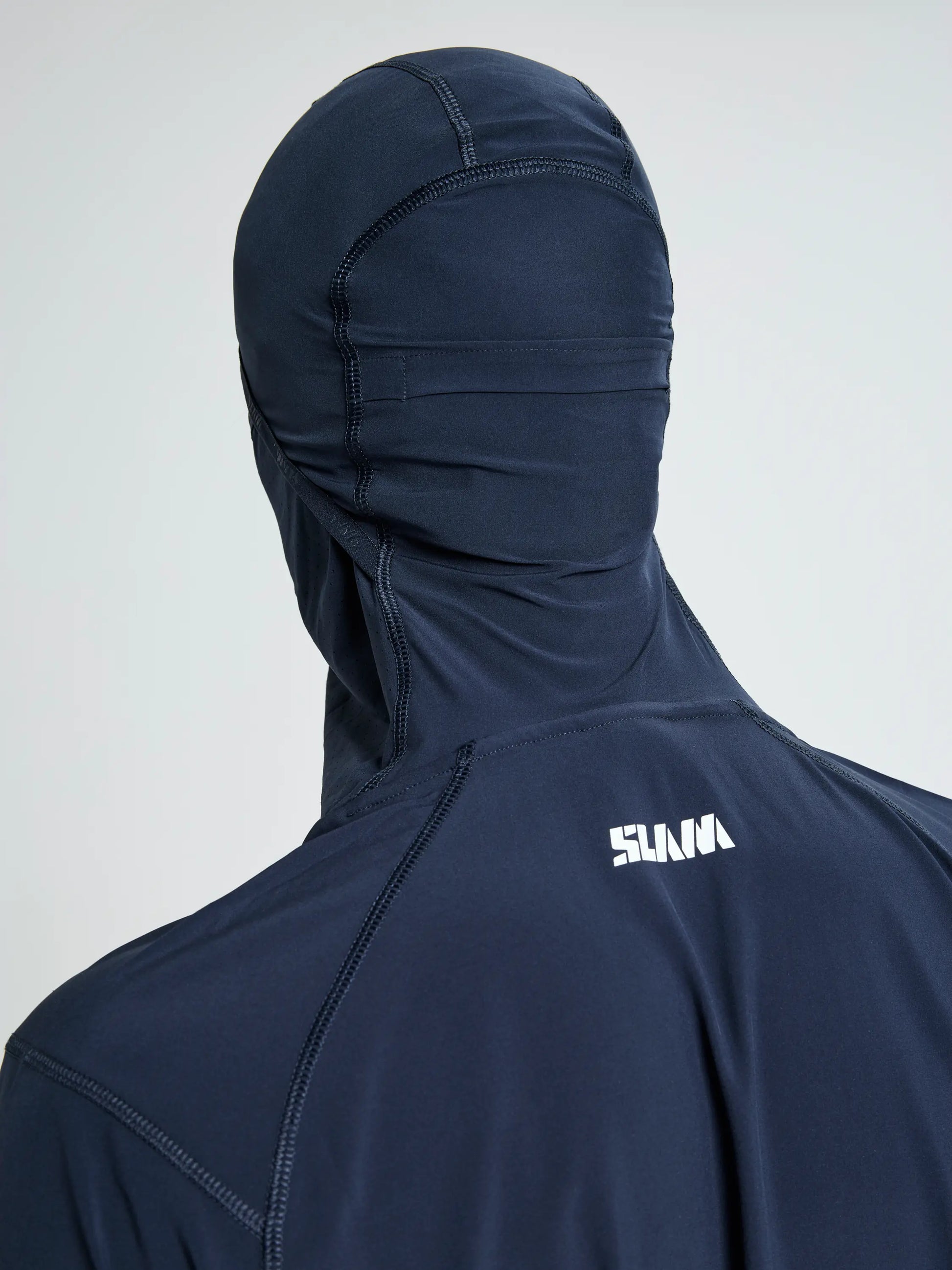 A person facing away wears the SLAM OD TECH HOODED T-SHIRT: a fitted, dark navy top with hood covering the head and neck, crafted from recycled stretch fabric, featuring visible seams and a white "SUNA" logo on the upper back.