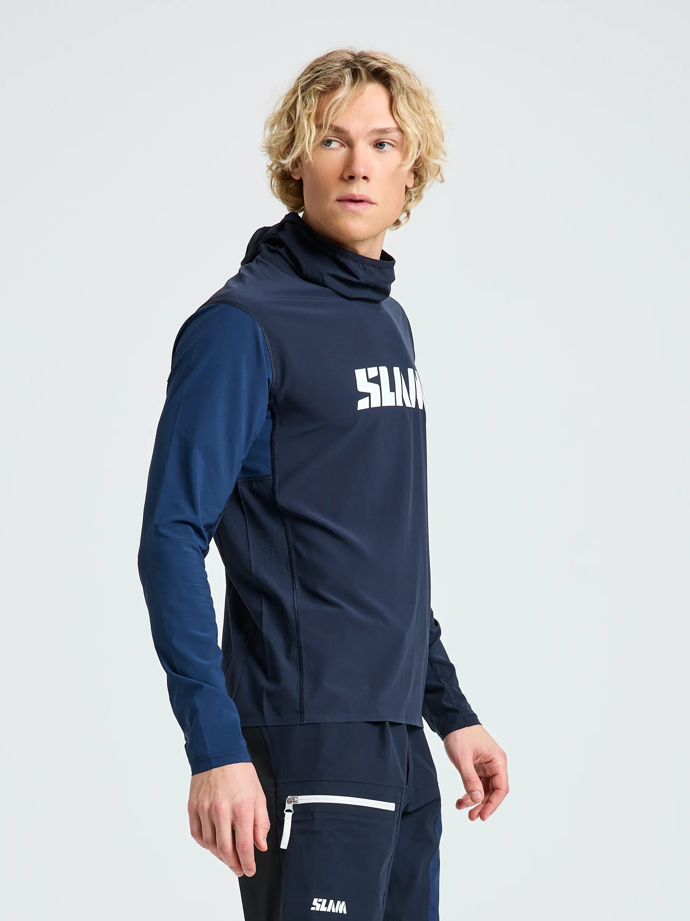 A person with wavy blond hair wears the SLAM OD TECH HOODED T-SHIRT in navy, featuring a white "SLAM" logo on the chest, paired with matching navy pants. They face slightly right against a plain light background.