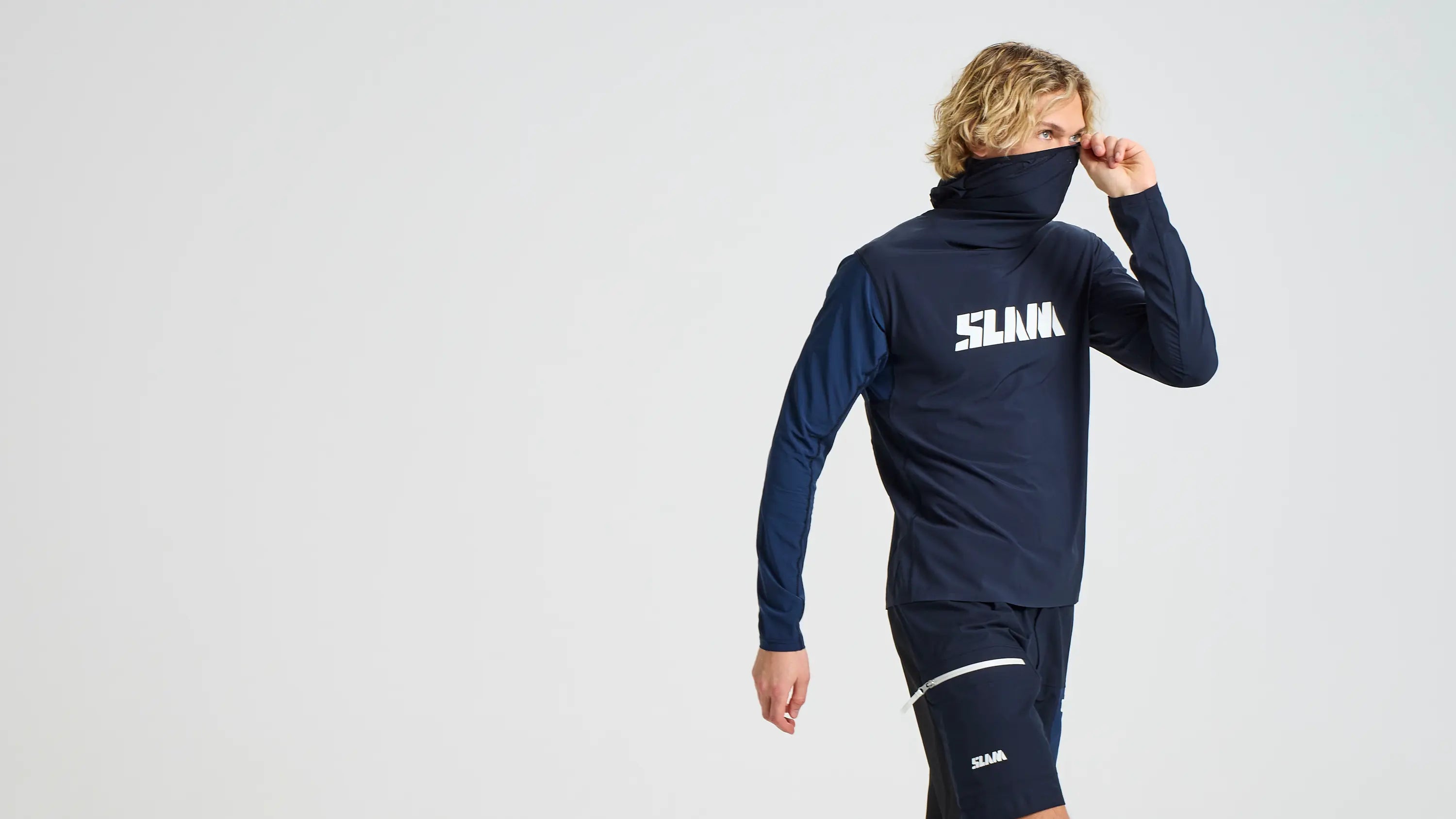 A blonde-haired person wears the SLAM OD TECH HOODED T-SHIRT and matching shorts, both in navy blue recycled stretch fabric with "SLAM" print, partially hiding their face with the collar against a plain light background.