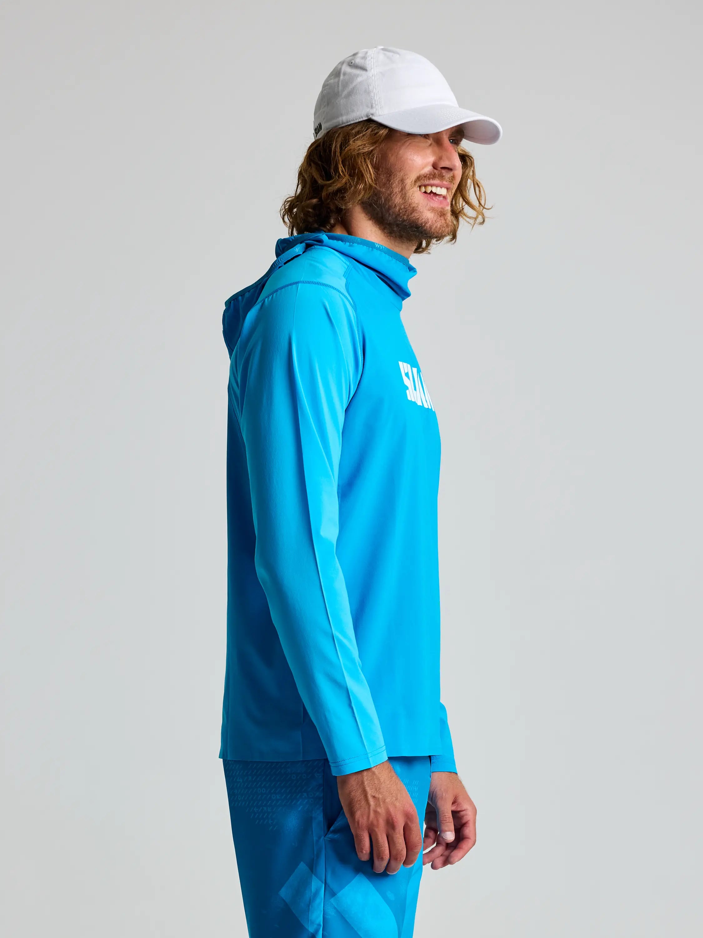 A man with curly hair, in a white cap, stands sideways in a bright blue OD TECH HOODED T-SHIRT by SLAM. Made from recycled stretch fabric and featuring antibacterial properties, "SALOMON" is written on the shirt against a plain gray background.