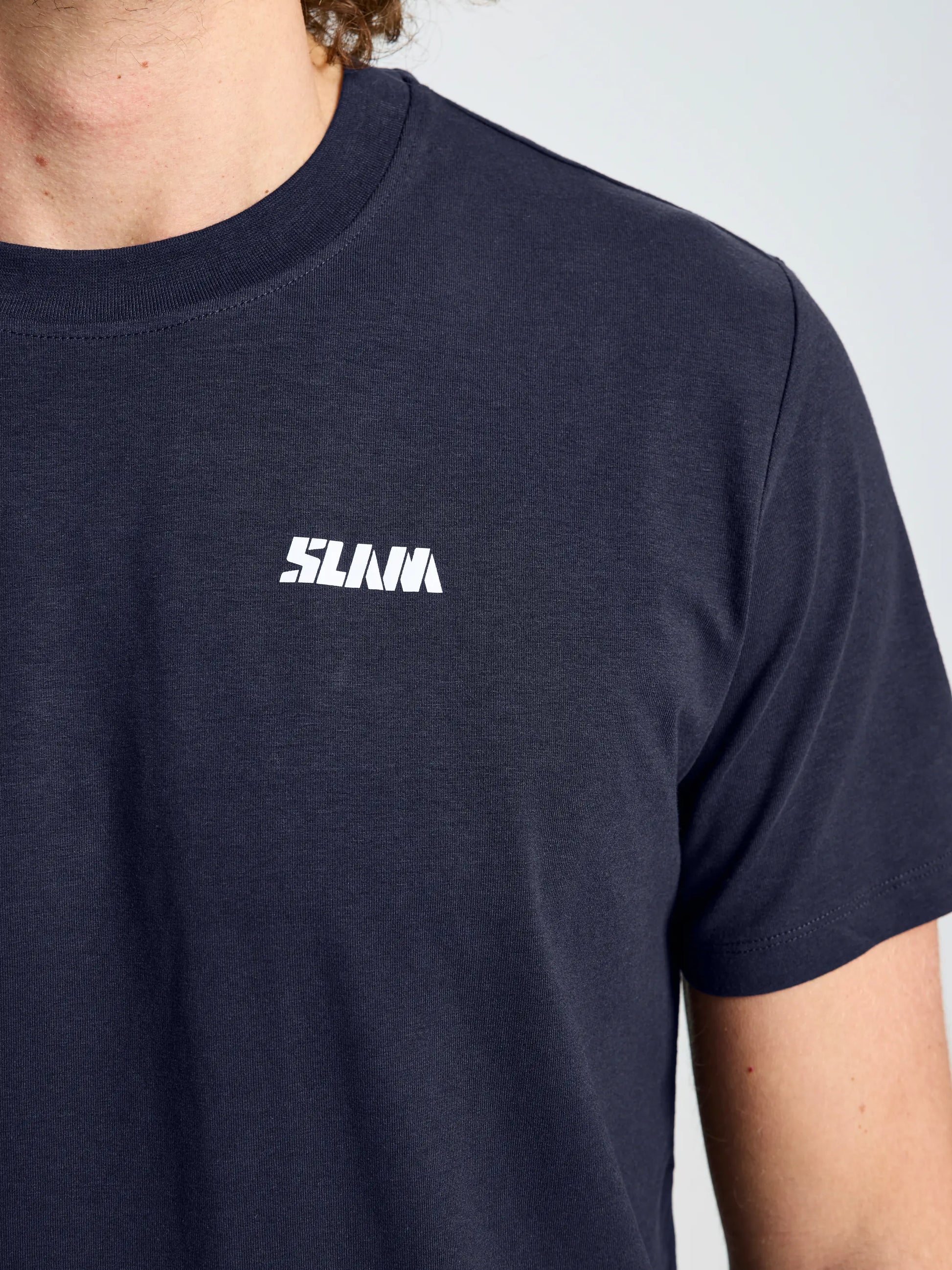 Close-up of a person wearing the SLAM GRAPHIC T-SHIRT SS in dark navy blue, crafted from soft cotton-bamboo blend, with "SLAVA" in white uppercase letters on the left chest. Only part of neck, shoulder, and upper arm are shown.