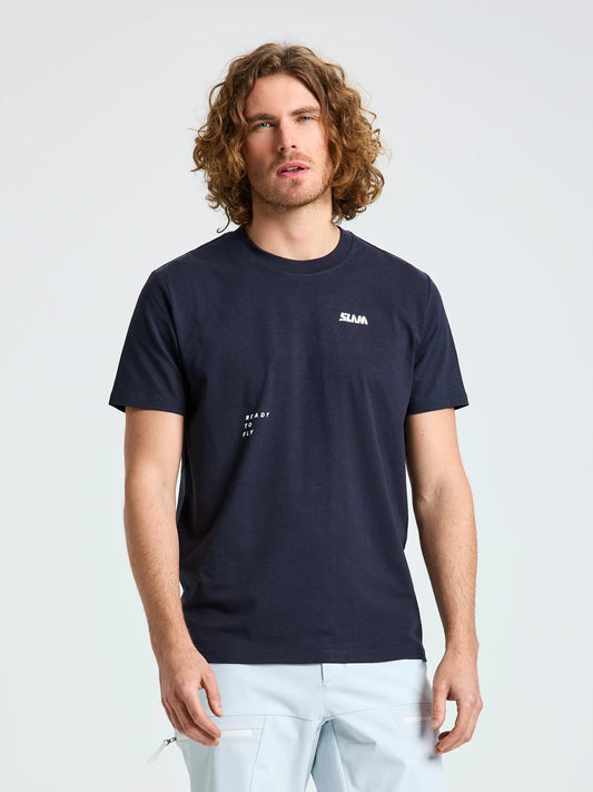 A man with curly hair and a beard wears the SLAM GRAPHIC T-SHIRT SS in navy blue, made from soft cotton and bamboo, featuring small white text, paired with light blue pants. He stands facing forward against a plain light background.