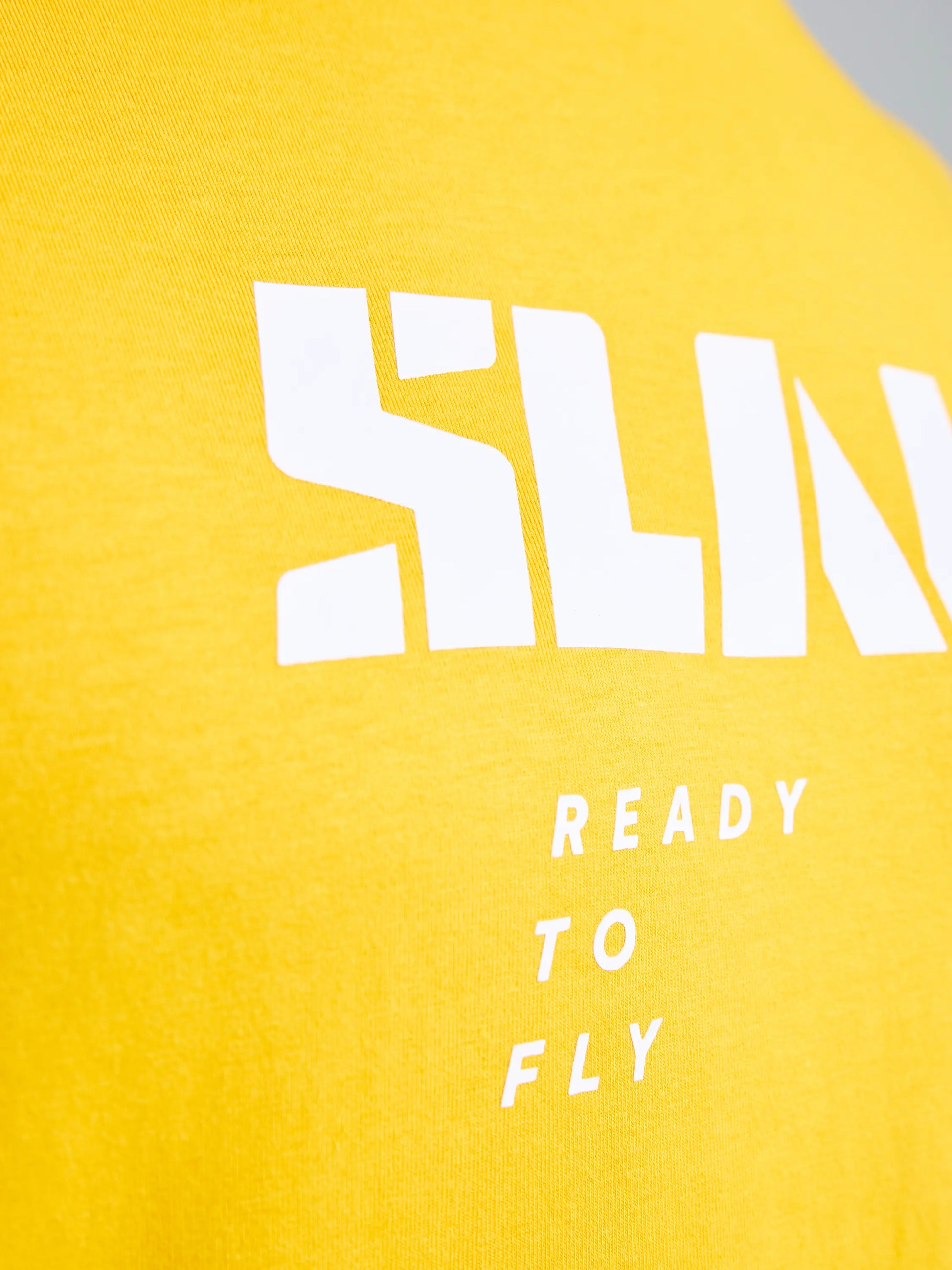 Close-up of the SLAM GRAPHIC T-SHIRT SS in yellow, crafted from soft cotton and bamboo fabric. Bold white letters are partially visible at the top with "READY TO FLY" in smaller white text beneath.