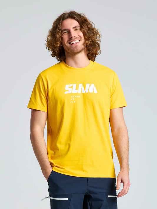 A curly-haired man stands smiling in a bright yellow SLAM GRAPHIC T-SHIRT SS made with cotton and bamboo, featuring bold white "SLNJA READY TO FLY" text, paired with dark pants, against a plain light background.