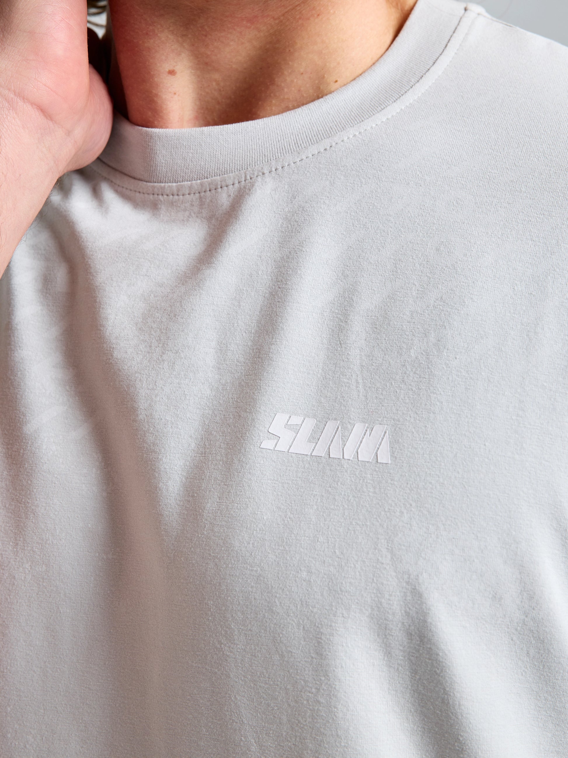 A person wears the SLAM WIND MAP T-SHIRT in light gray bamboo jersey, featuring "SLAM" in white on the chest, and touches the side of their neck.
