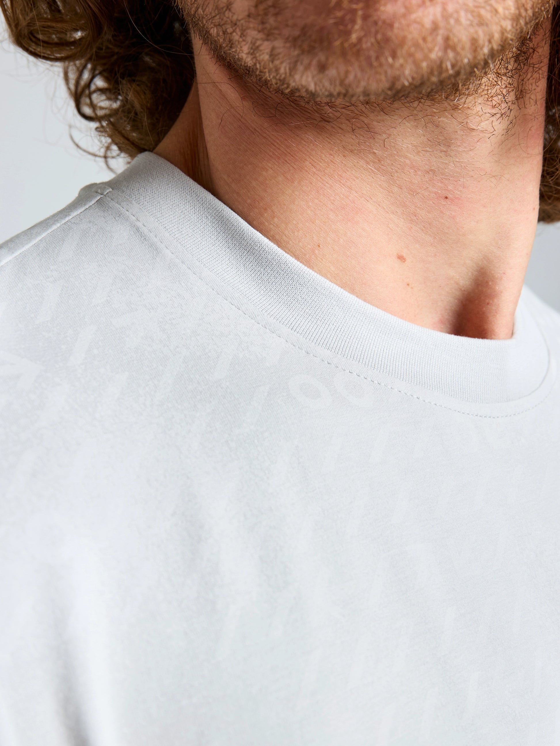 Close-up of a man's lower face, neck, and shoulder wearing the SLAM WIND MAP T-SHIRT in light grey bamboo jersey. The lightweight tee has a subtle, barely visible pattern with only part of his curly hair, beard, and mustache in view.