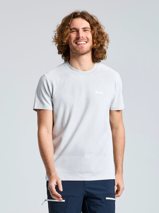 A person with shoulder-length curly hair smiles while standing against a plain light background, wearing the SLAM WIND MAP T-SHIRT in light gray bamboo jersey and dark pants with zipper pockets.