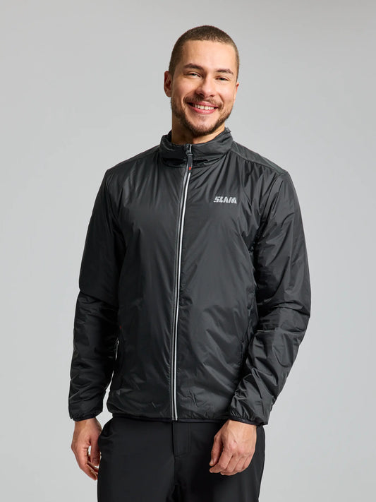 A person is smiling while wearing a compressible, graphite-colored PRO ALPHA LIGHT JACKET for men by SLAM, which features a small logo on the chest. They are standing against a plain, light gray background.