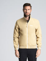 A man stands facing forward against a plain light gray background, wearing the SLAM ALTURA DOCK JACKET in CORDA BEIGE with white accents and a small white logo on the left chest. He maintains a neutral expression.