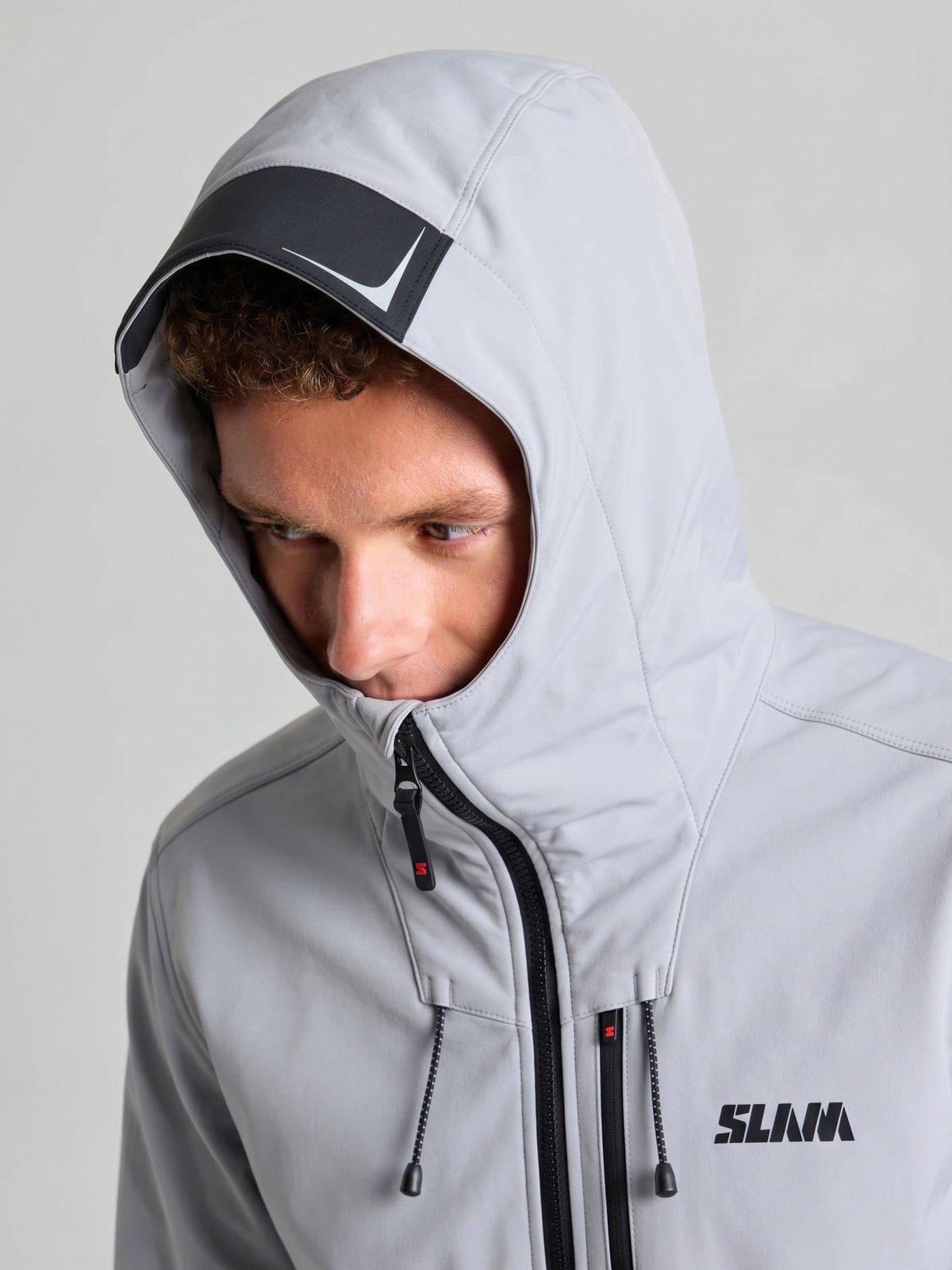 A person wearing a light gray SLAM SOFTSHELL HOODIE with black zippers and drawstrings. The hood is up, covering their head, and the jacket has “SLAM” printed on the chest.
