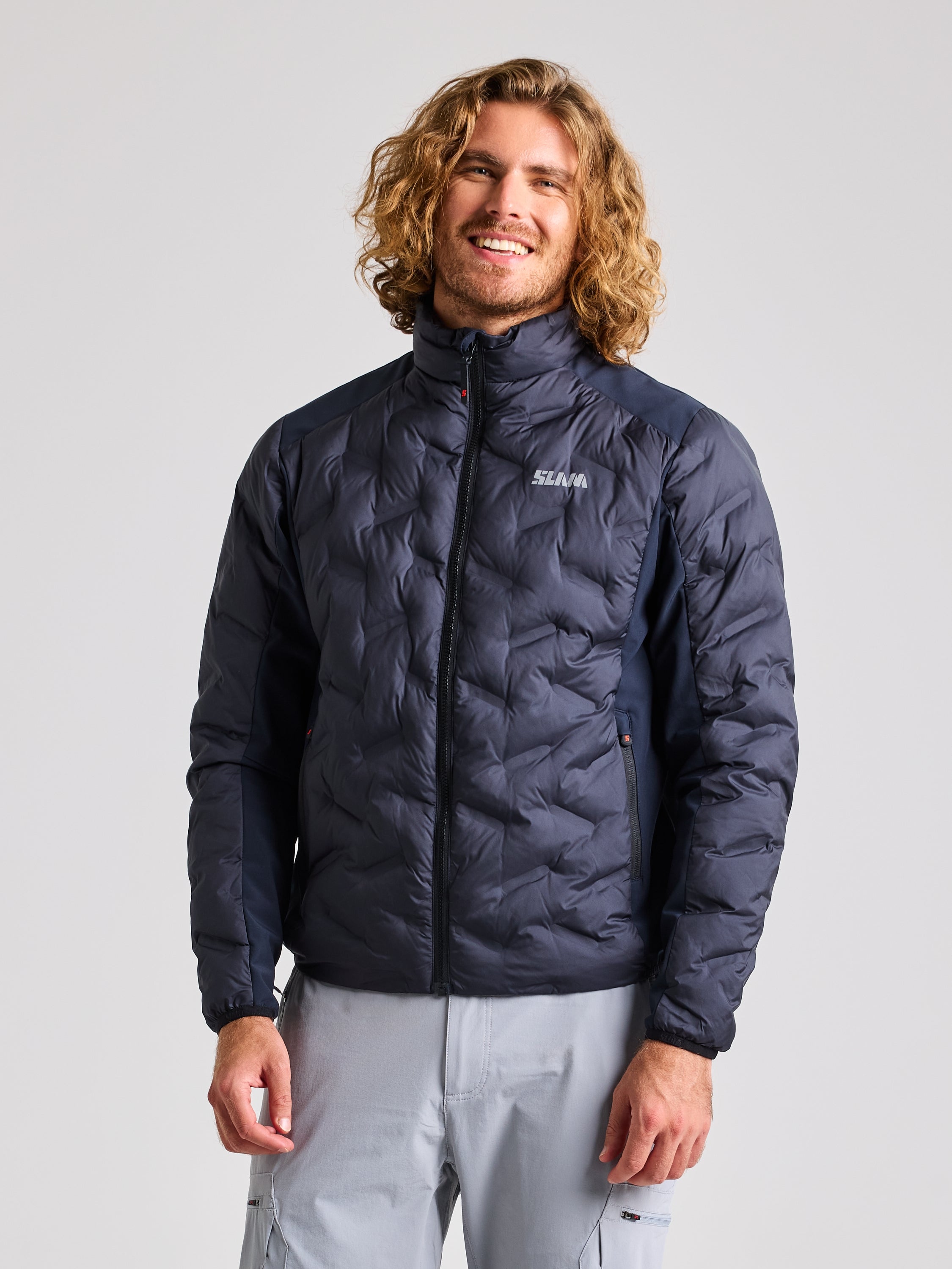 A person with long hair is wearing a dark navy HYBRID SOFTSHELL JACKET by SLAM, featuring the word "Selva," paired with light-colored pants. This stylish quilted jacket not only looks great but also offers excellent protection. The person stands against a plain background, smiling warmly.
