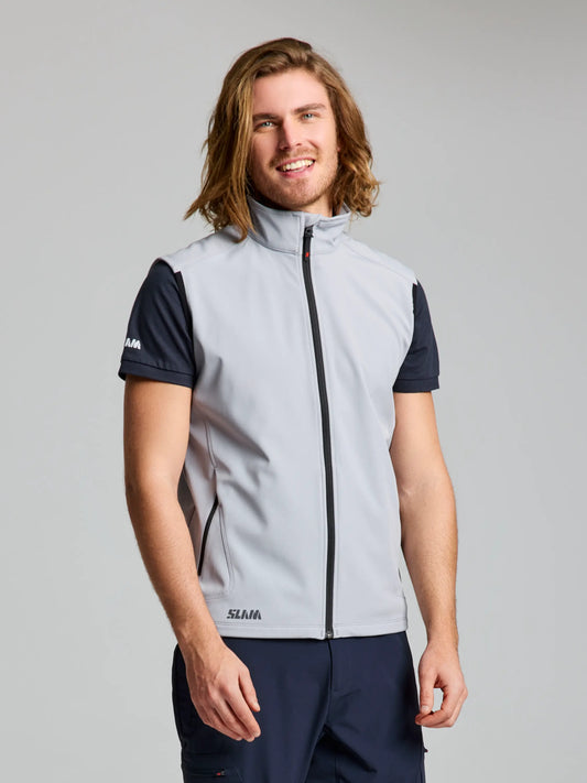 A man with long hair is standing and smiling, wearing the AIRYUM-colored ACTIVE SOFTSHELL VEST from SLAM over a short-sleeve black shirt and dark pants. Tailored for athletes, this vest provides protection against wind and water. The background is plain and neutral in color.