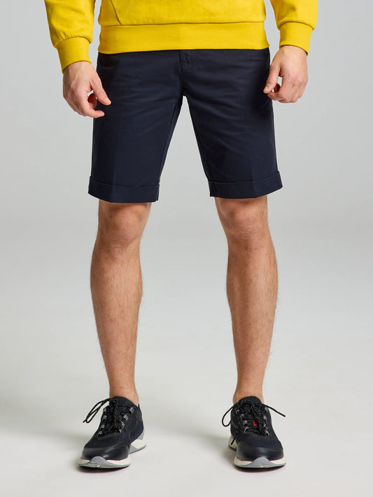 A person in a yellow sweatshirt and men's DECK LIGHT CHINO SHORT from SLAM is standing against a plain background, their DARK NAVY shorts and black athletic shoes creating a subtle nautical look. The shot artfully highlights their legs and torso, capturing the essence of casual elegance.