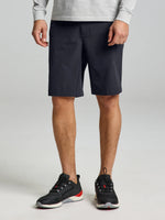 A person stands against a plain light background, shown from the shoulders down, wearing SLAM TECH CHINO SHORTS in a dark color styled at knee length with a stretch nylon fabric, paired with a light long-sleeve shirt and black athletic shoes featuring white soles.