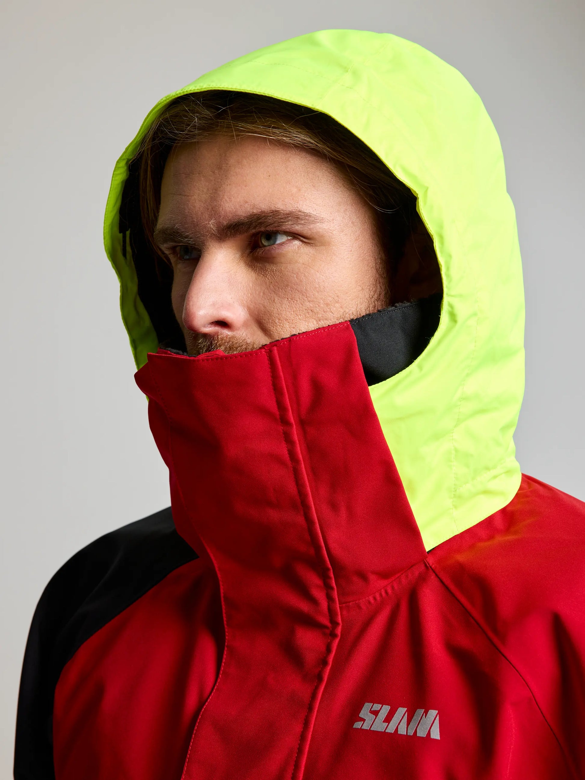 A person wears the SLAM PRO COASTAL JKT—a red and black waterproof sailing jacket with a high collar and neon yellow hood, featuring “SLAM” on the chest. They are looking to the side.