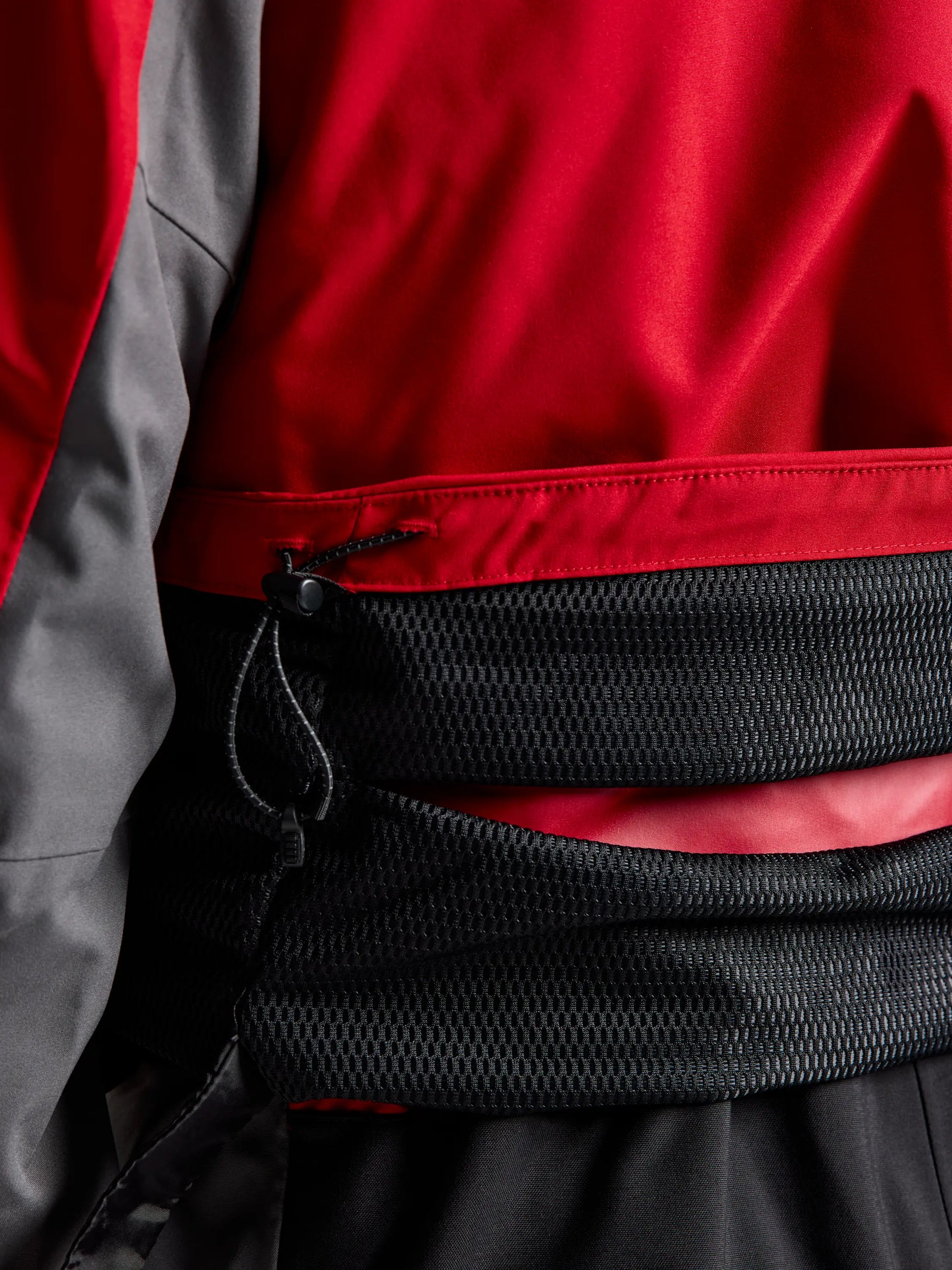 A close-up of a person wearing the SLAM PRO COASTAL JKT, a red and grey sailing jacket with a black mesh waist belt secured by an adjustable cord.