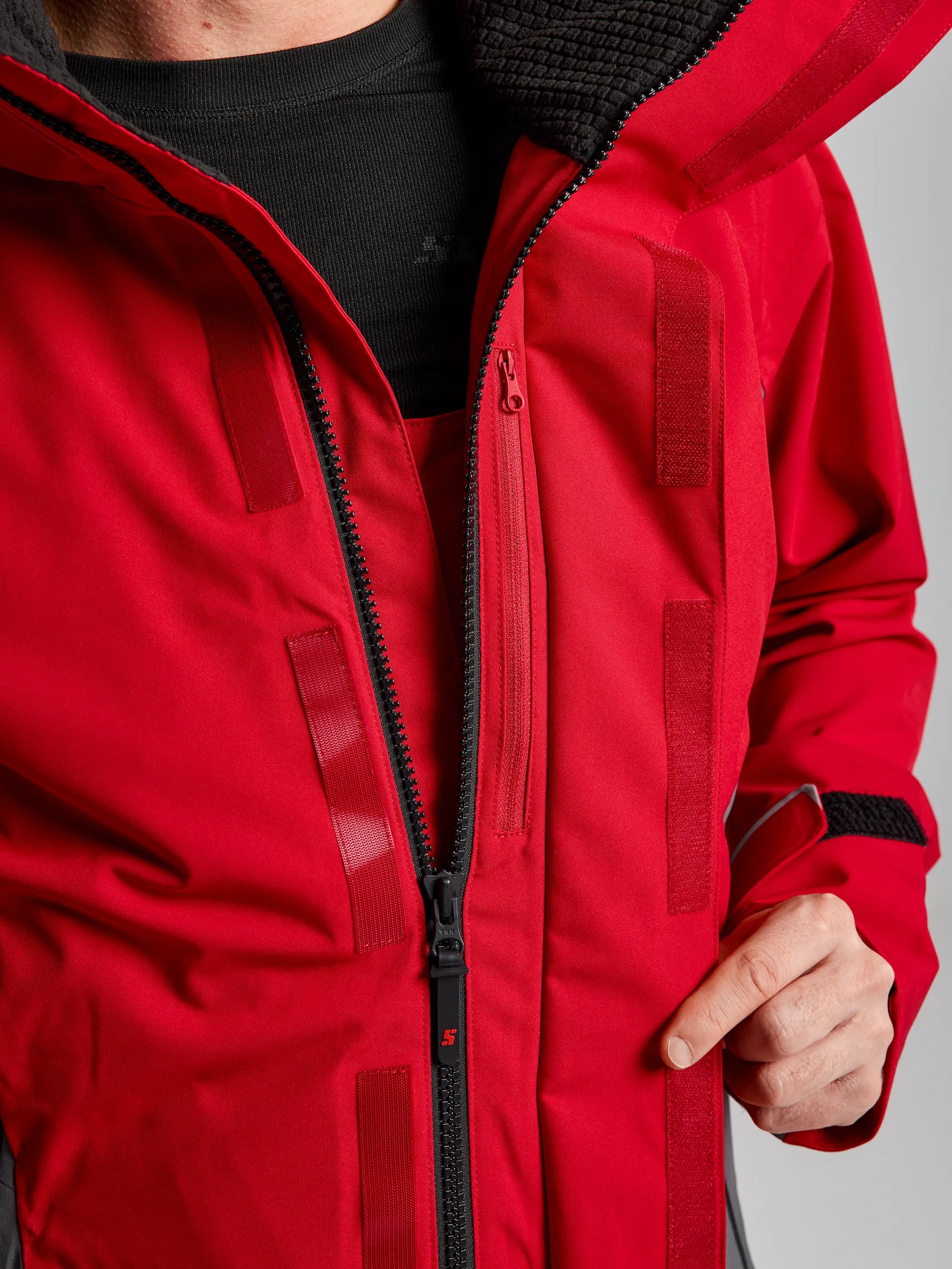 A person wearing the SLAM PRO COASTAL JKT, a red waterproof jacket with black zippers and velcro fastenings, is shown unzipping the front to reveal a black inner shirt. The photo highlights the torso and hand, with no face visible.