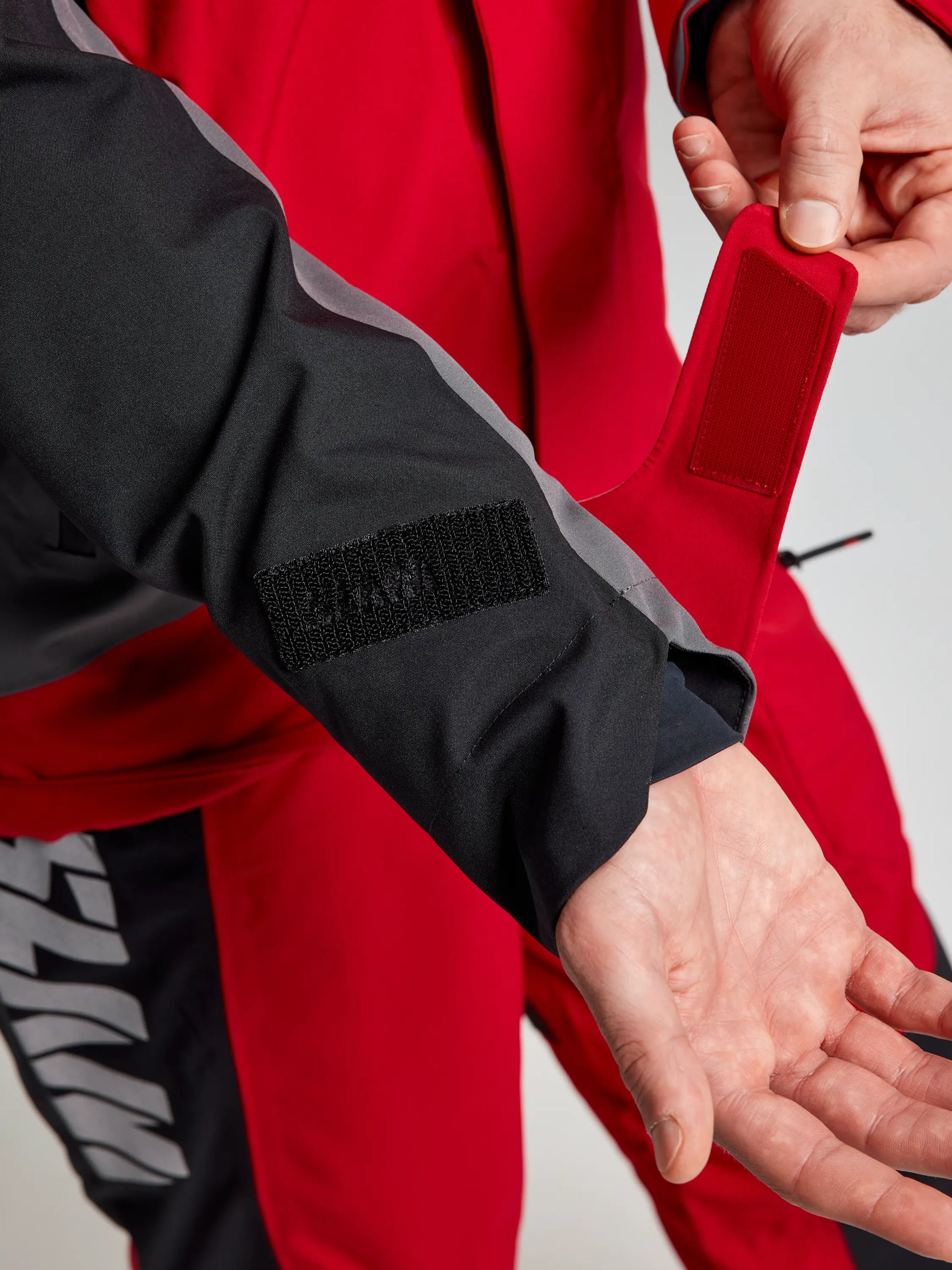 A person adjusts the red and black Velcro cuff on the sleeve of a SLAM PRO COASTAL JKT, revealing part of their open palm. Both the jacket and pants display bold white text.
