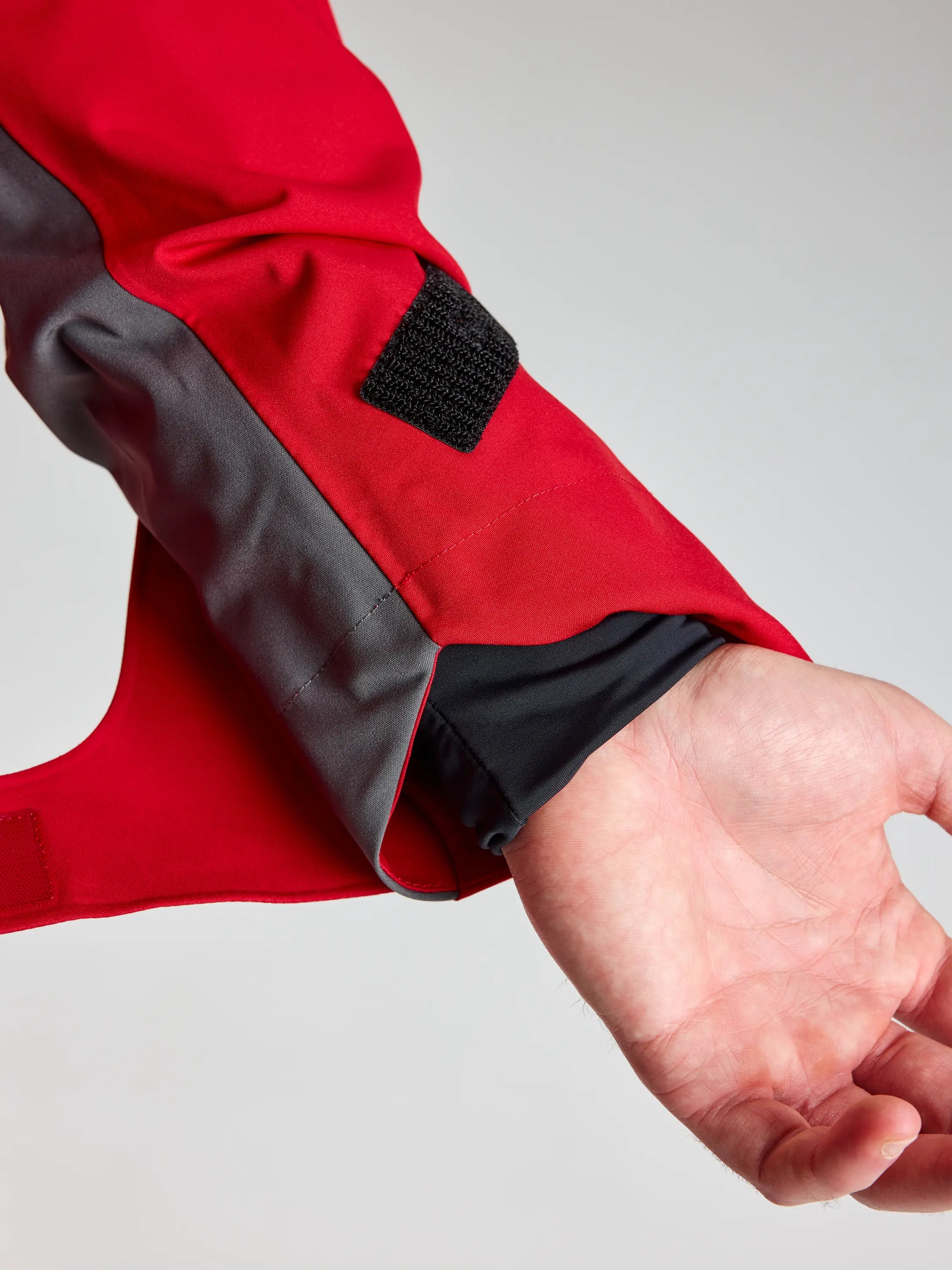 A close-up of a person's wrist and hand wearing the SLAM PRO COASTAL JKT in red and gray, featuring an adjustable black Velcro sleeve strap. The jacket is partially open to show a black inner layer.