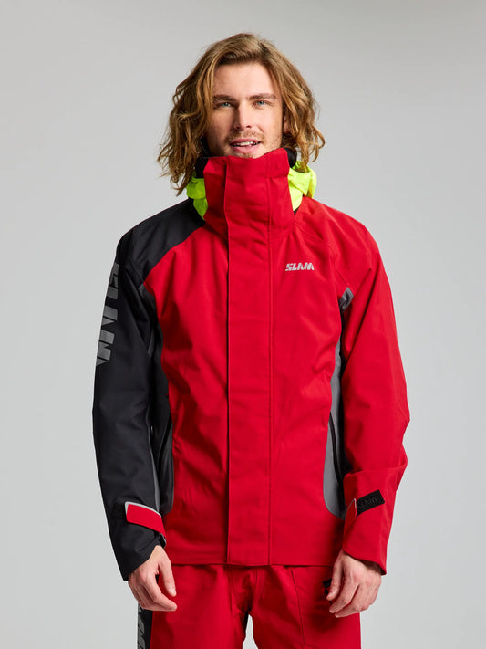 A man with long hair wears a SLAM PRO COASTAL JKT in red and black, featuring a high collar and neon yellow hood, as he faces the camera against a plain light grey background.
