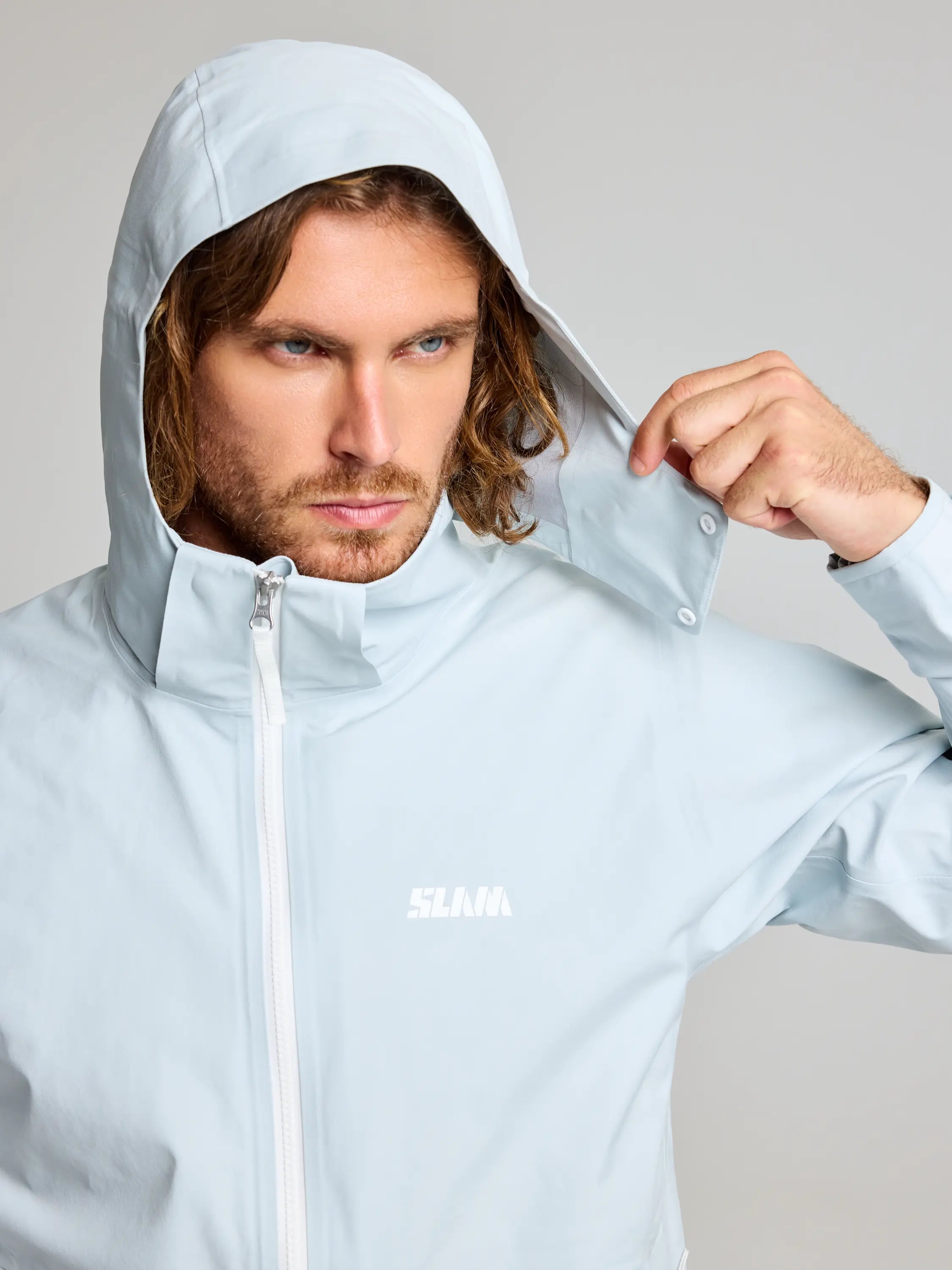 A person with long hair wears a SLAM FOIL HOODED JACKET in light blue, pulling its breathable hood over their head with one hand. The jacket includes a white zipper, chest logo, and is waterproof. The background is plain light gray.