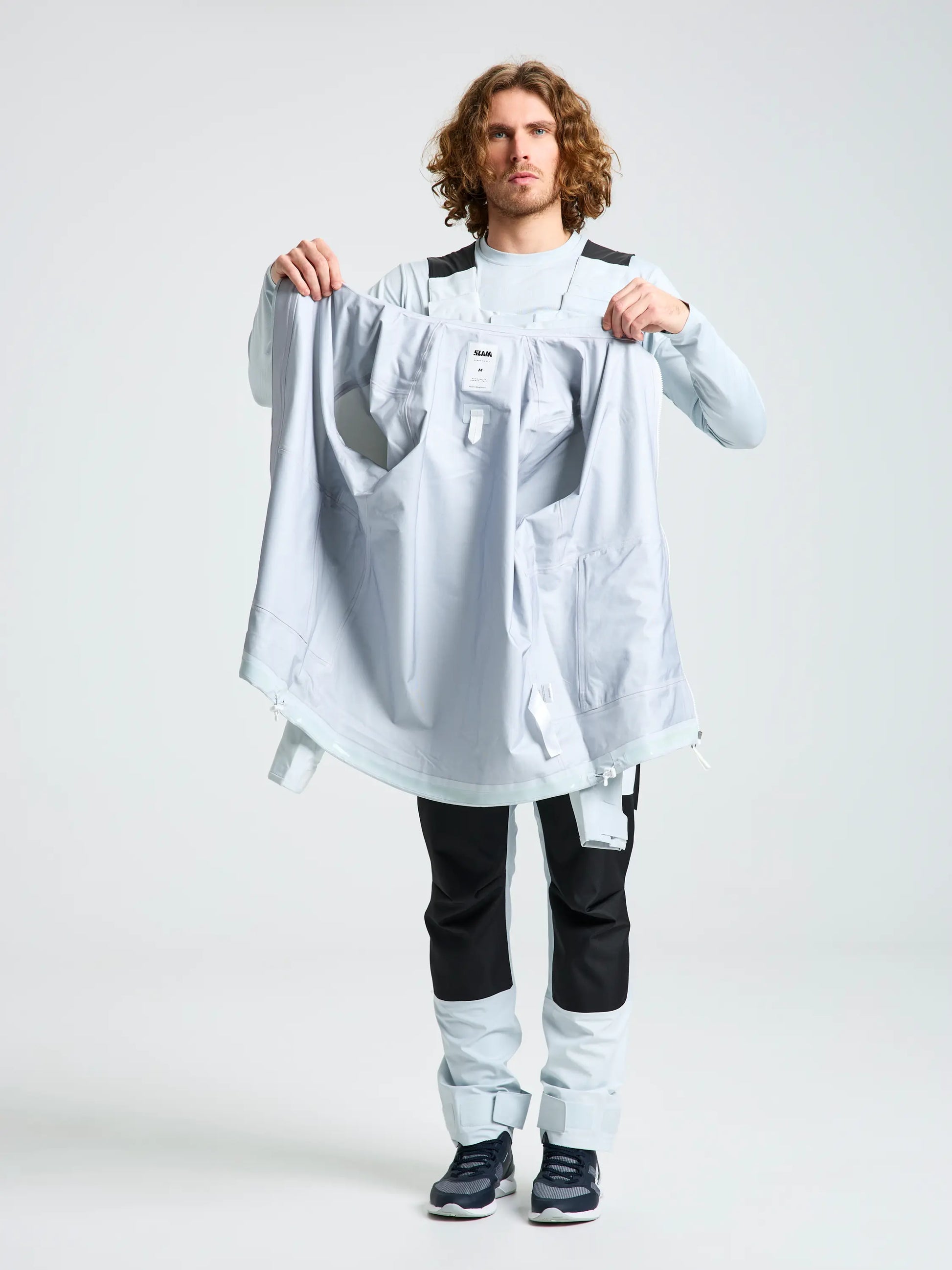 A person with shoulder-length curly hair faces forward, holding open the SLAM OD HOODED JACKET in light gray to show its interior, paired with light outerwear pants featuring black accents and dark sneakers.
