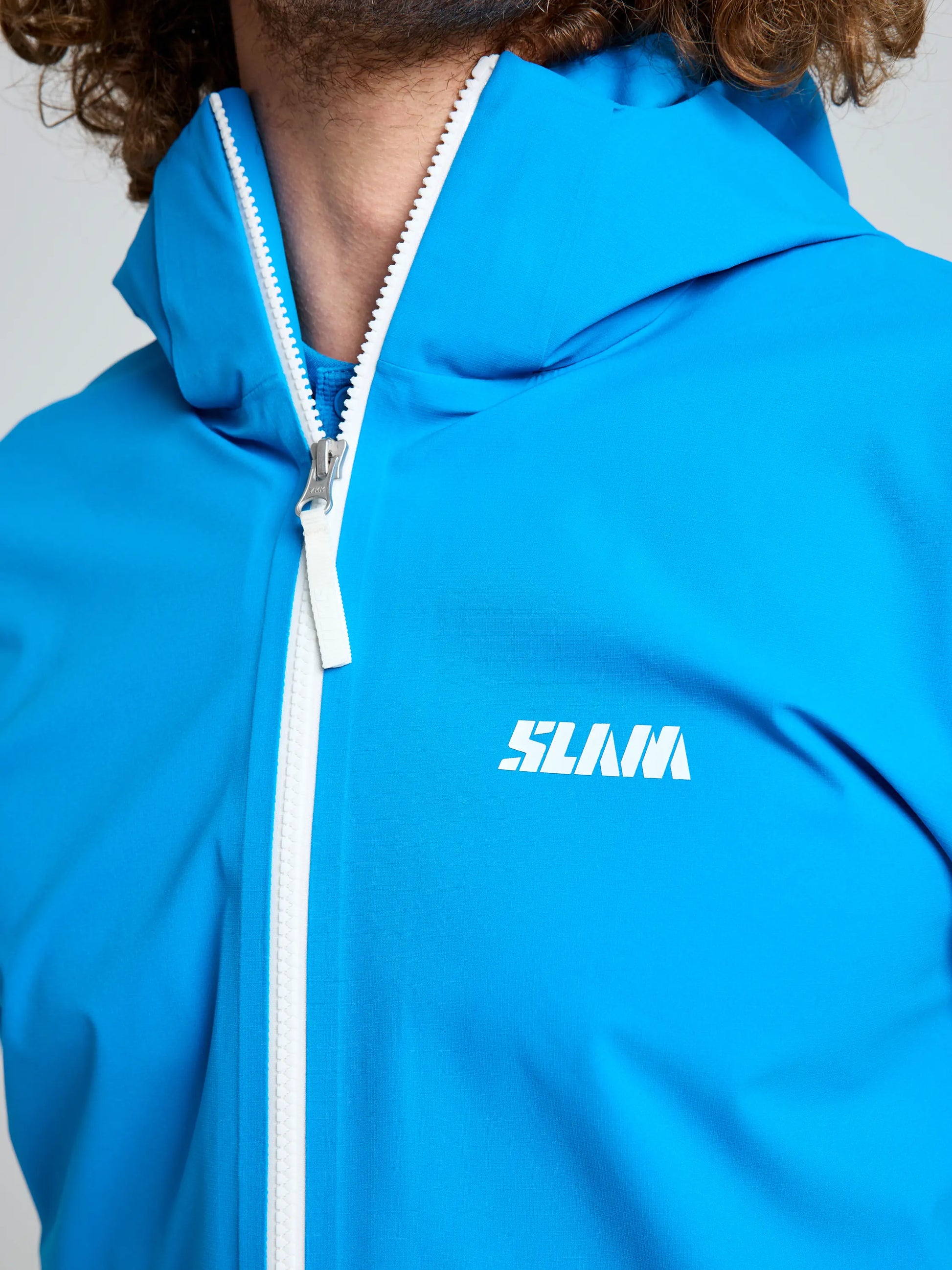 A close-up of a person in a bright blue SLAM OD HOODED JACKET with a white zipper and "SLAM" printed in white on the left chest, partially showing their curly brown hair and face.