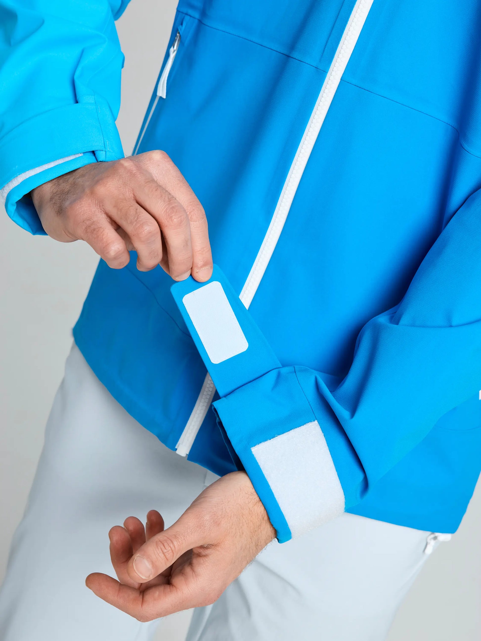 A person adjusts a rectangular white patch on the sleeve of a SLAM OD HOODED JACKET in bright blue, featuring a white zipper and Velcro cuff. Only their hands and part of their arms and torso are visible.
