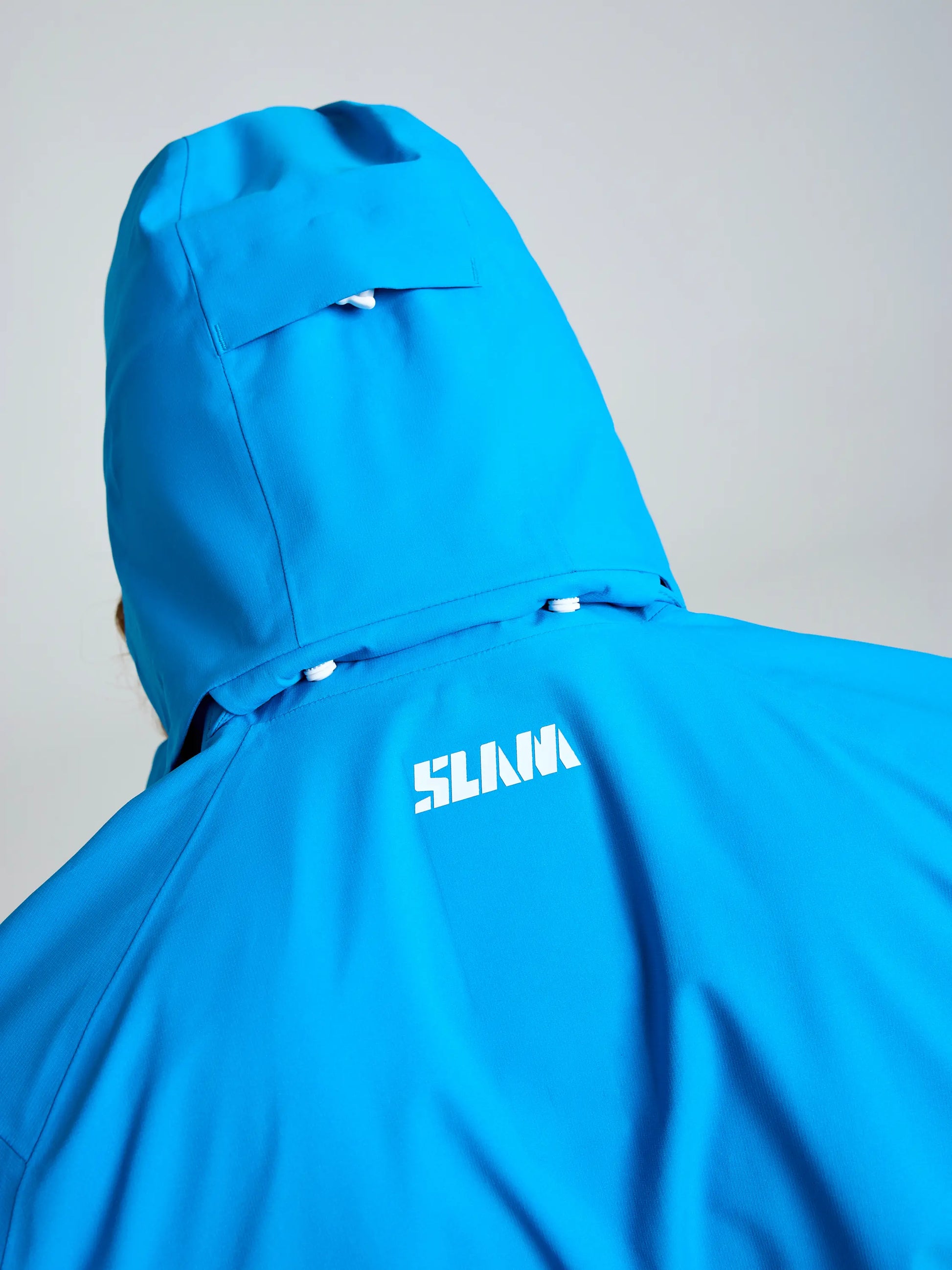 A close-up of the back of the bright blue OD HOODED JACKET by SLAM, featuring "SLAM" printed in white on the upper back and showcasing its hood and shoulder details against a plain background.
