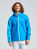 A man with medium-length curly hair smiles while wearing the SLAM OD HOODED JACKET, a bright blue sailing jacket with white zippers, and light-colored pants against a plain background.