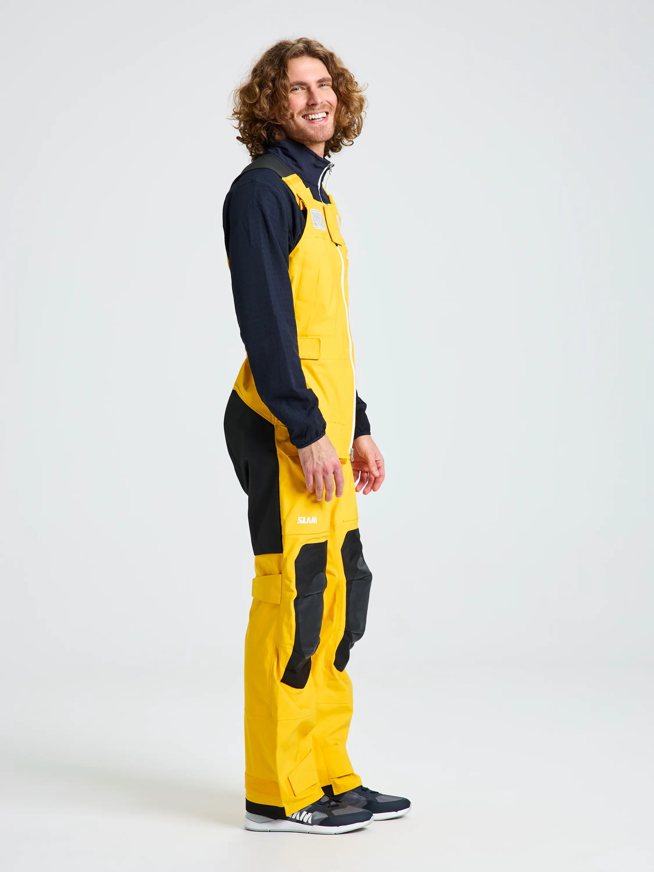 A person with curly hair stands sideways against a plain light background, wearing the SLAM OCEAN BIB—yellow and black technical jumpsuit with zippered pockets—and black sneakers.